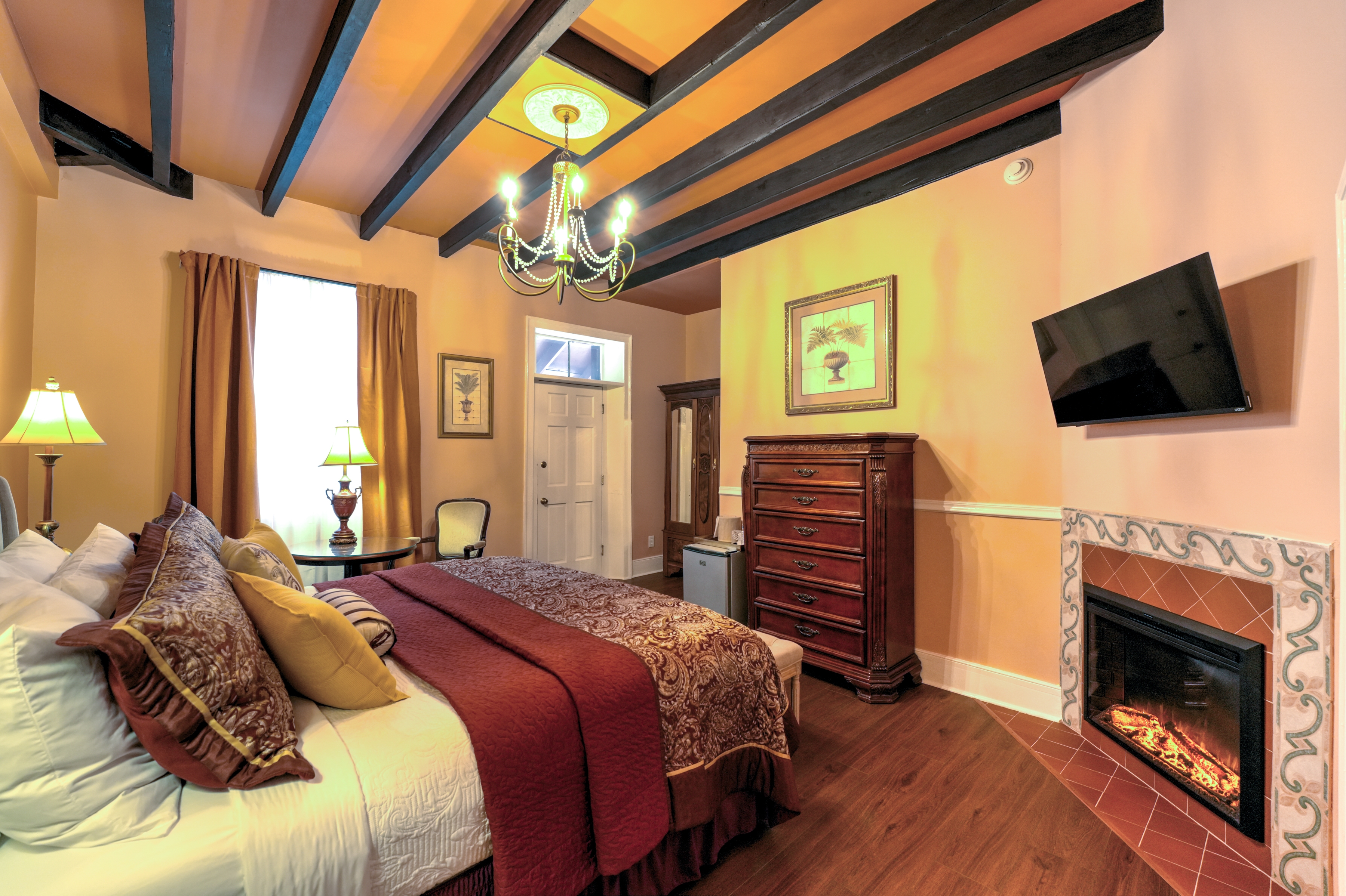 Spacious bedroom with a king bed with an upholstered headboard, antique armchairs and dresser on opposite wall, and at the far end of the room a door leading to the balcony, Fireplace and TV on one the corner wall