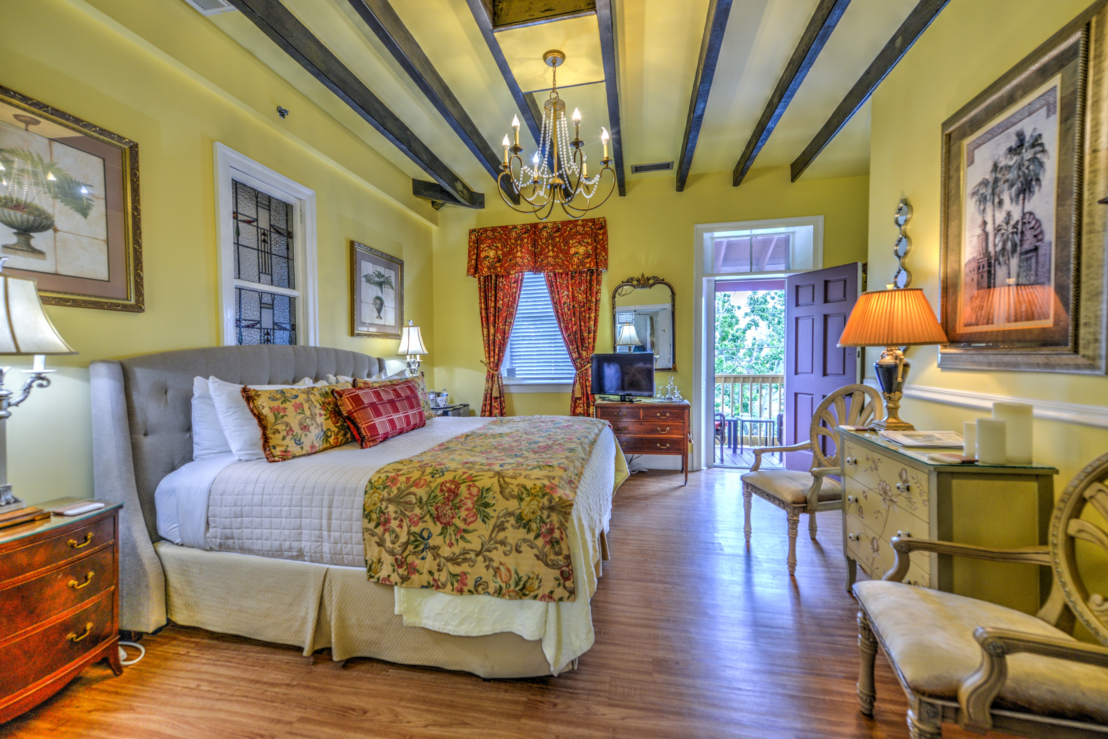 Spacious bedroom with a king bed with an upholstered headboard, antique armchairs and dresser on opposite wall, and at the far end of the room a door leading to the balcony