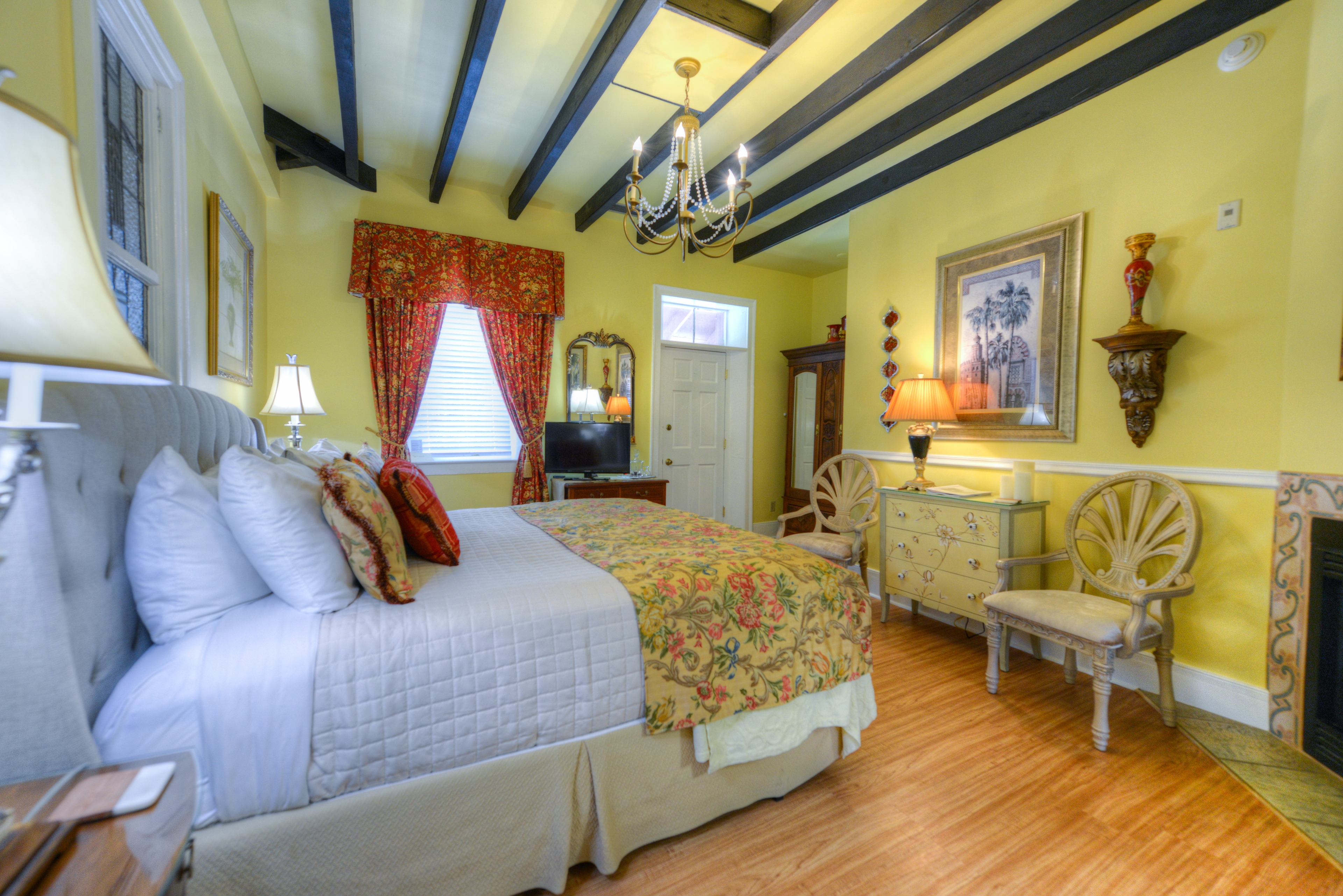 Elegant bedroom with king bed topped with decorative pillows, white sheets, and a floral spread.  The spacious room has a high, beam ceiling and is well appointed with antique furniture and artwork.