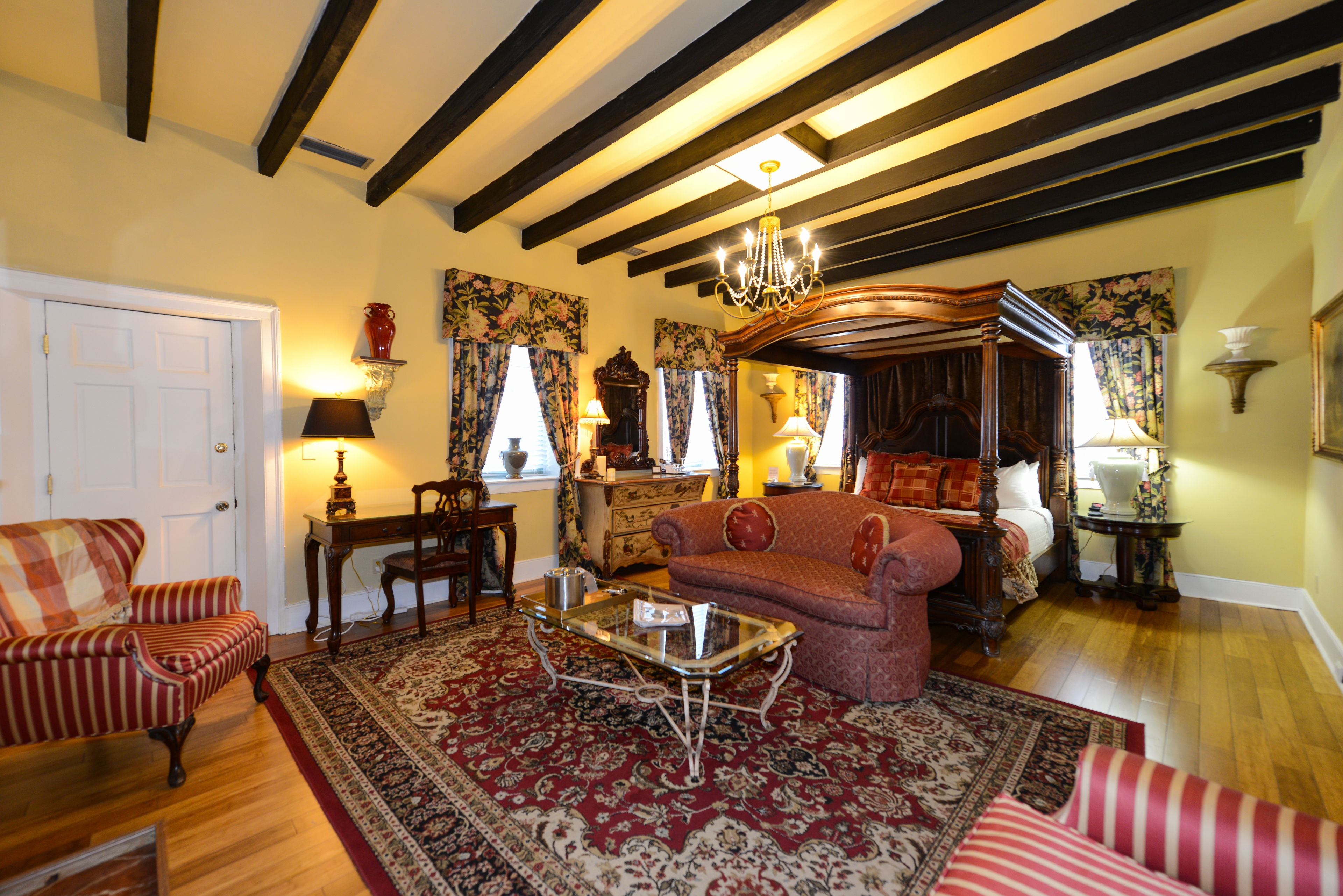 Romantic, luxurious guestroom with canopy four-poster king bed, beam ceiling, and sitting area with oriental area rug and upholstered loveseat and armchairs