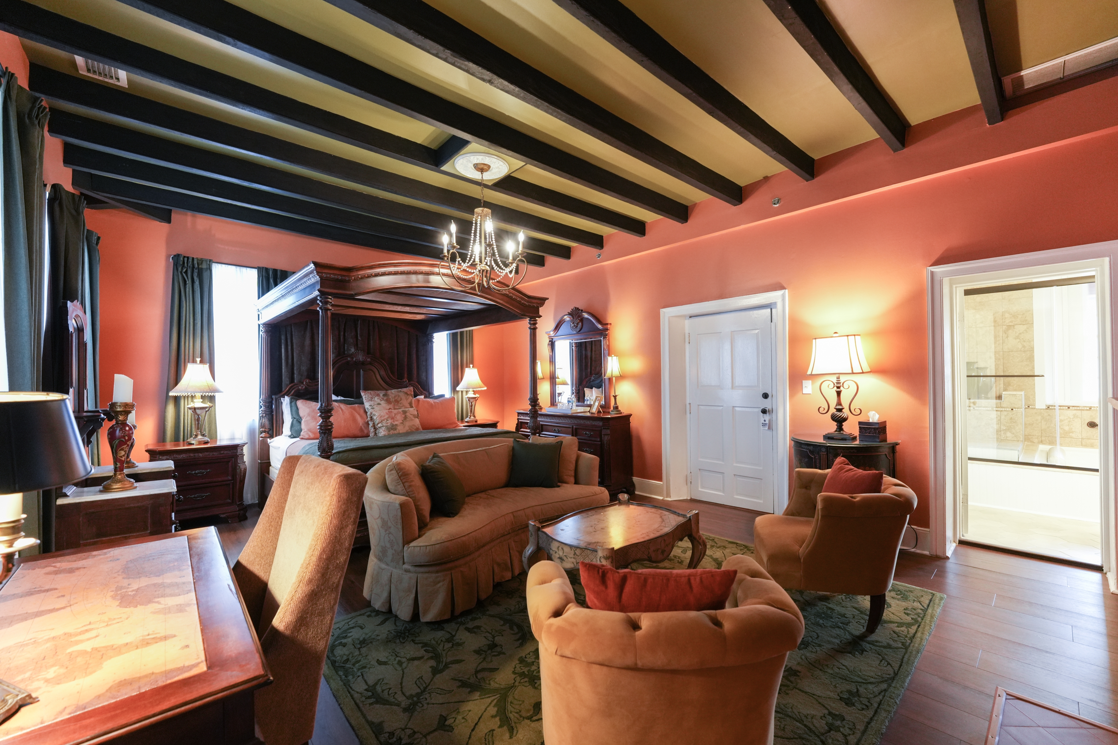 Romantic, luxurious guestroom with canopy four-poster king bed, beam ceiling, and sitting area with oriental area rug and upholstered loveseat and two chairs.  A doorway leads to the private bath.