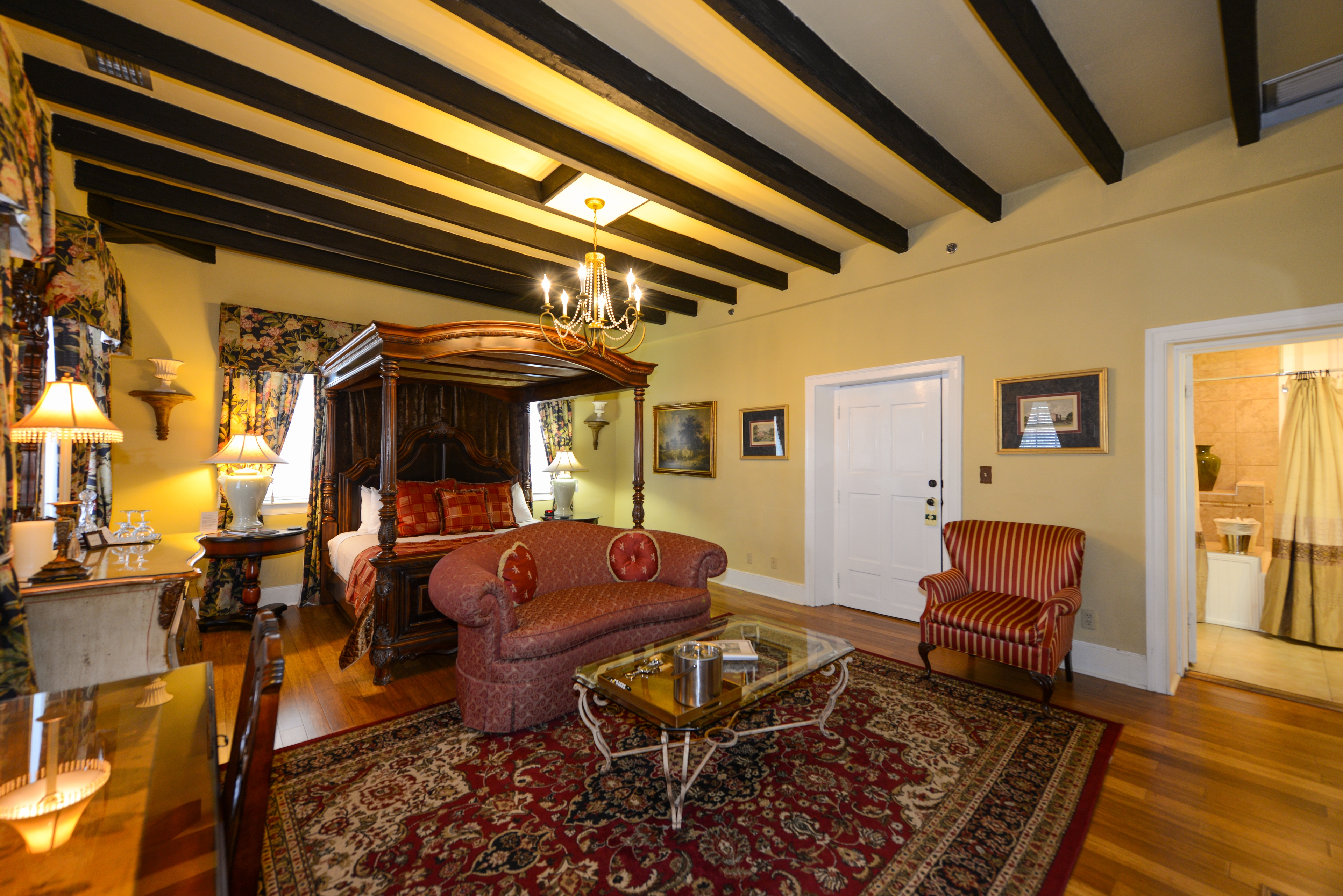 Romantic, luxurious guestroom with canopy four-poster king bed, beam ceiling, and sitting area with oriental area rug and upholstered loveseat and armchairs.  A doorway leads to the private bath.