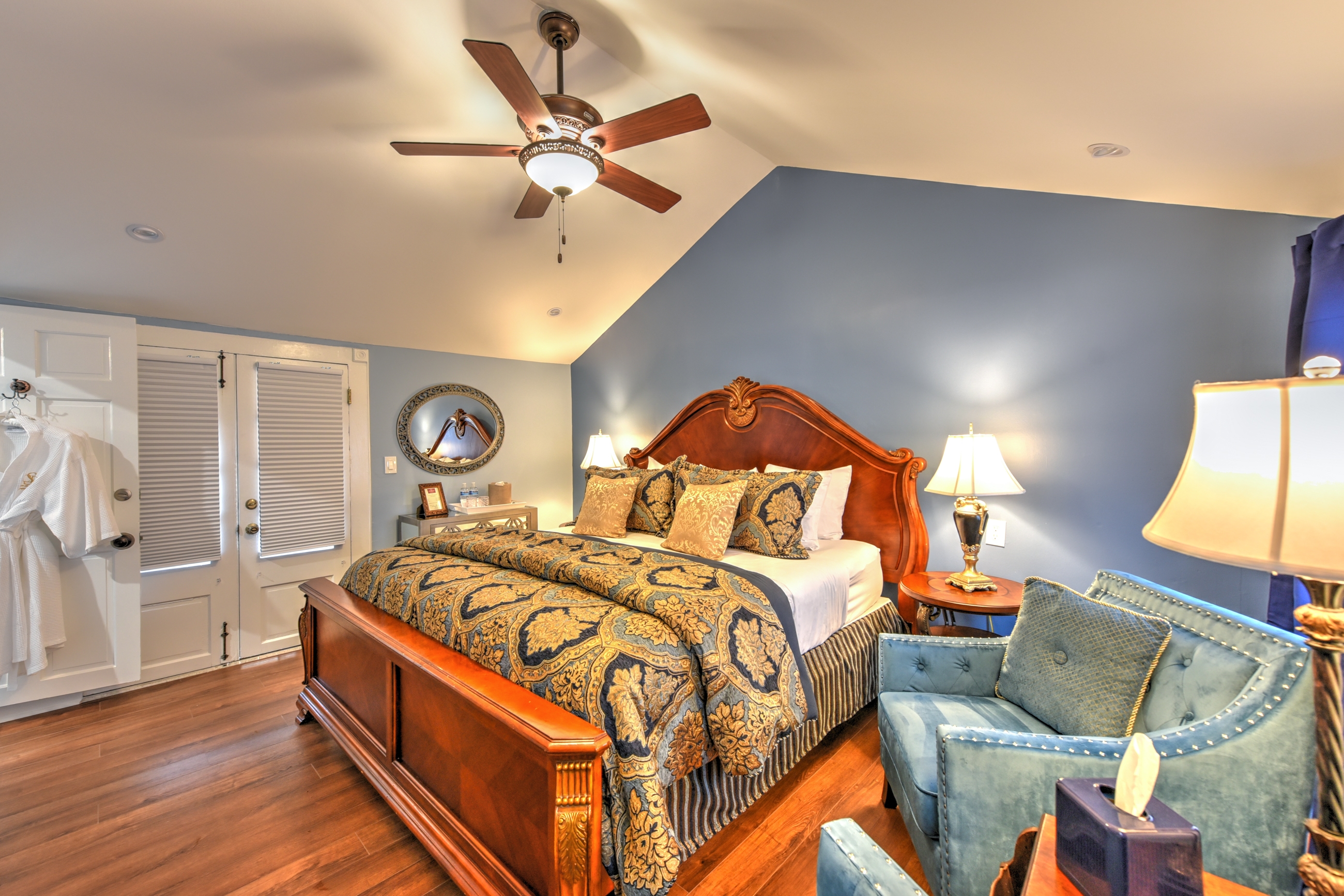 Bedroom with queen bed comfortably topped with blankets and decorative pillows. A flat-screen TV is mounted on the opposite wall, and two large windows let in natural light at the far end of the room.