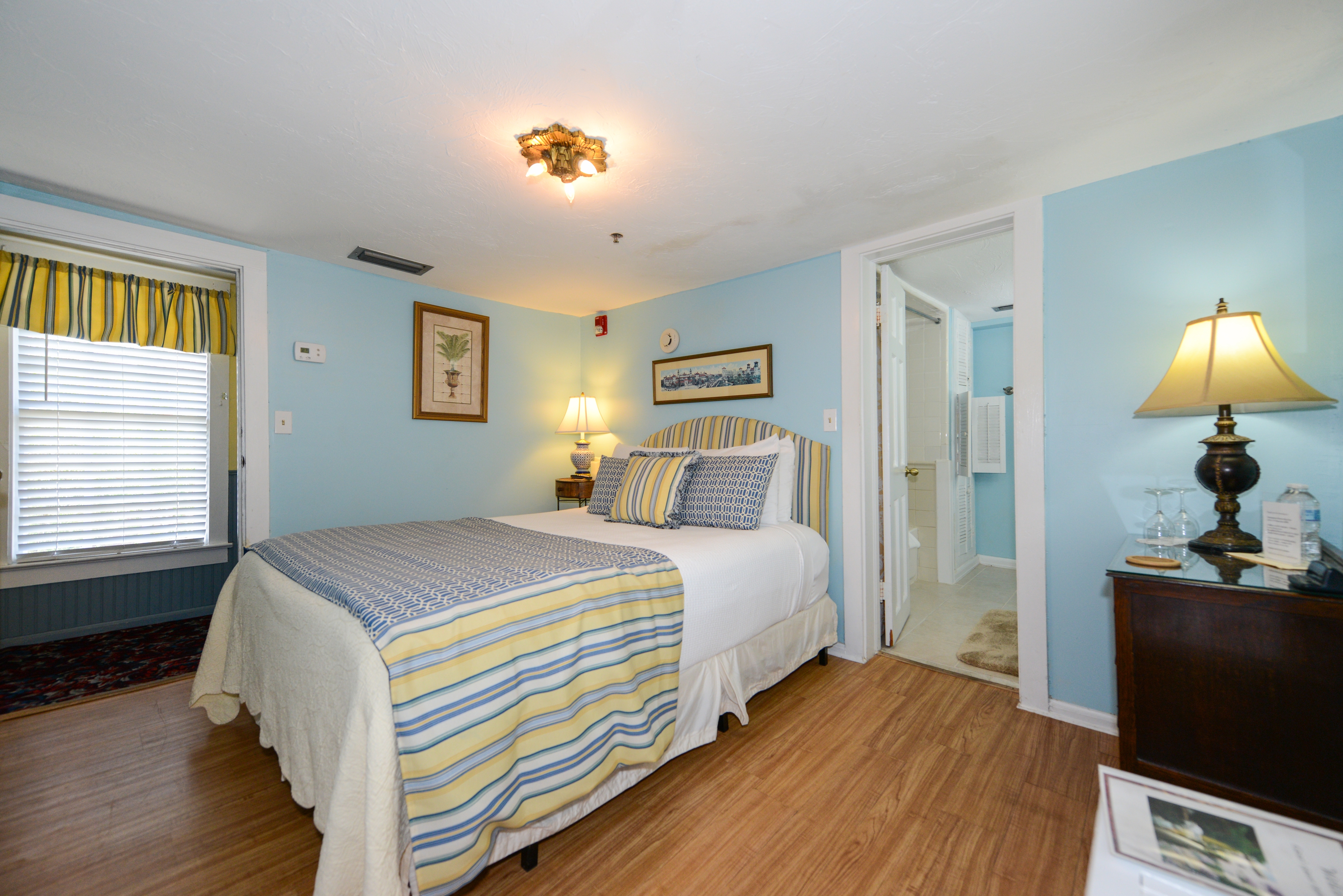 A queen bed with striped upholstered headboard with matching bedspread is the focal point of a bright, cheerful room.  A large window allows lots of natural light into the room.  Next to the bed is the doorway to the private bath, and on the other side of the doorway is a dark wooden dresser with an antique-style table lamp.