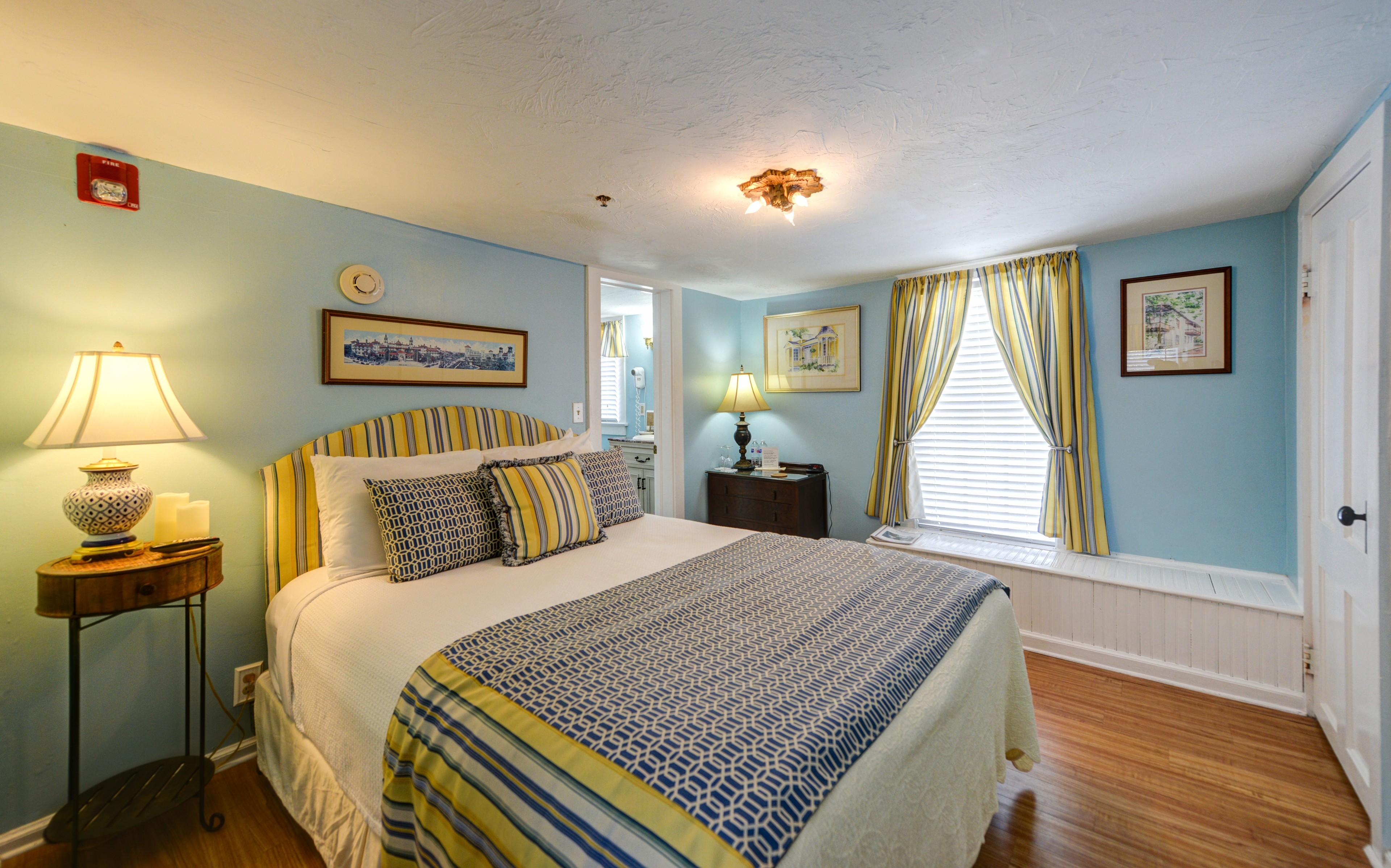 Charming bedroom decorated in light blue, yellow, white with a queen bed, sunny window with built-in window seat, and finished wood floors.