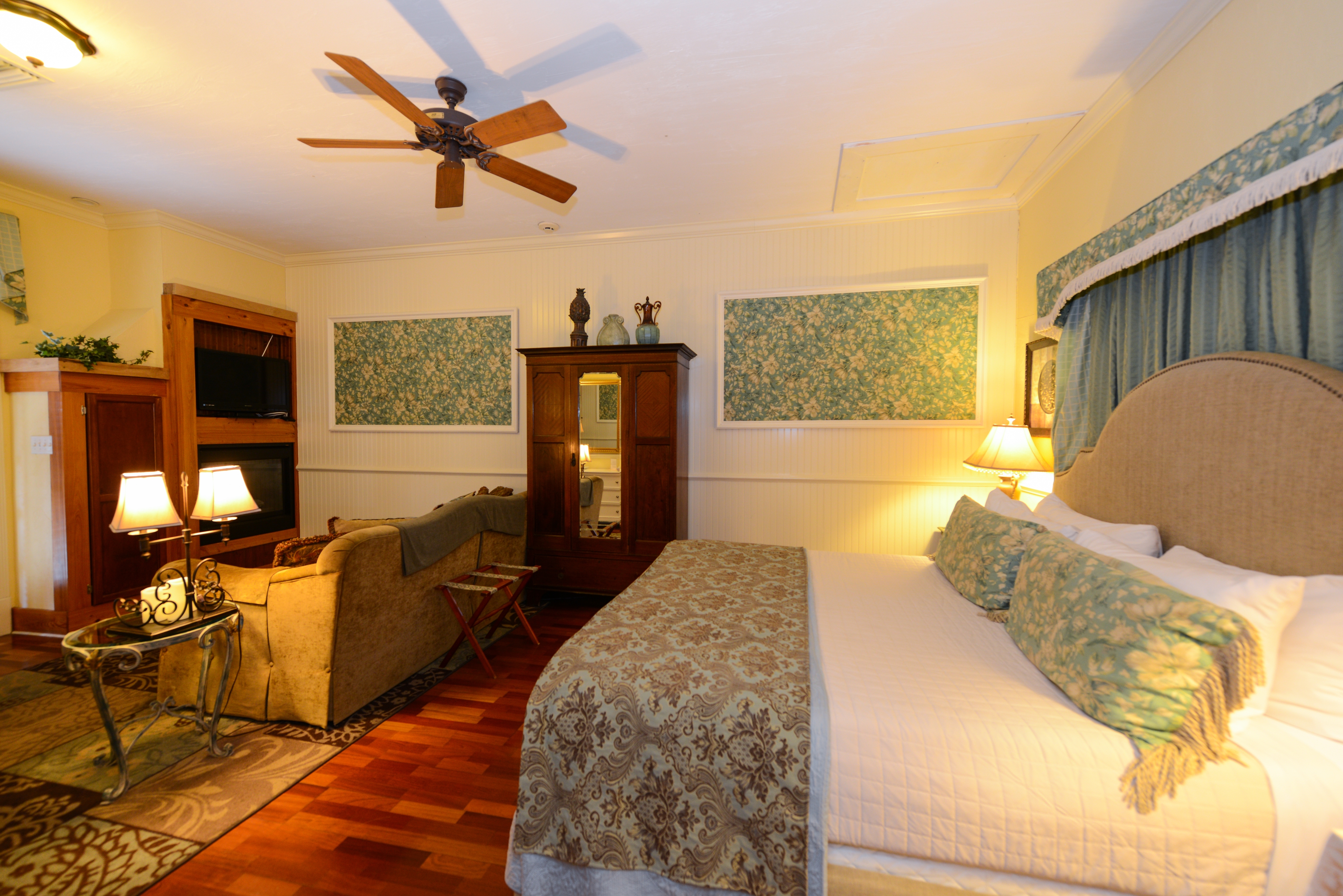 Spacious guestroom with king bed featuring a canopied headboard and topped with decorative bolster pillows.  On the other side of the room is a comfortable seating area with a sofa, fireplace and flat-screen TV (not shown).