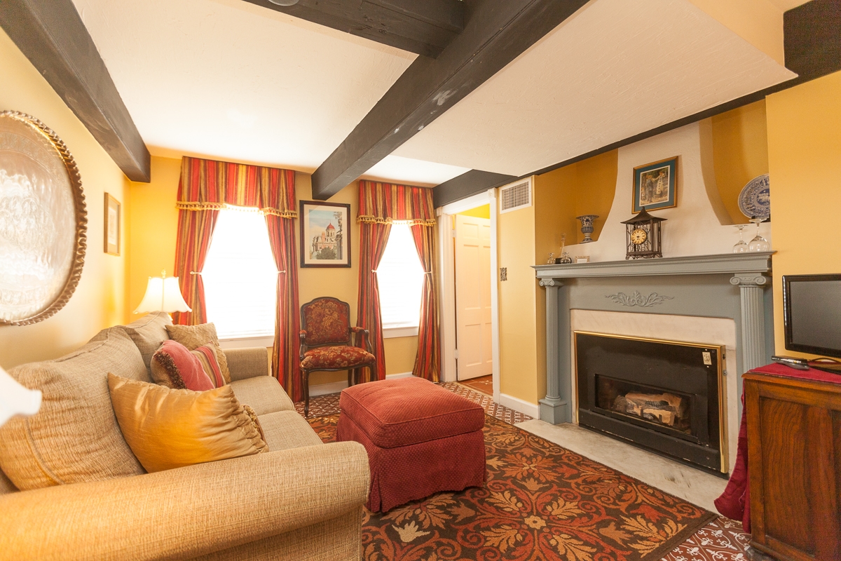 Sitting area decorated in shades of rich red and earth tones and comfortably appointed with sofa, armchairs and ottoman. Other features include two sunny windows with elegant drapes, beam ceiling, and fireplace with painted wood mantel.