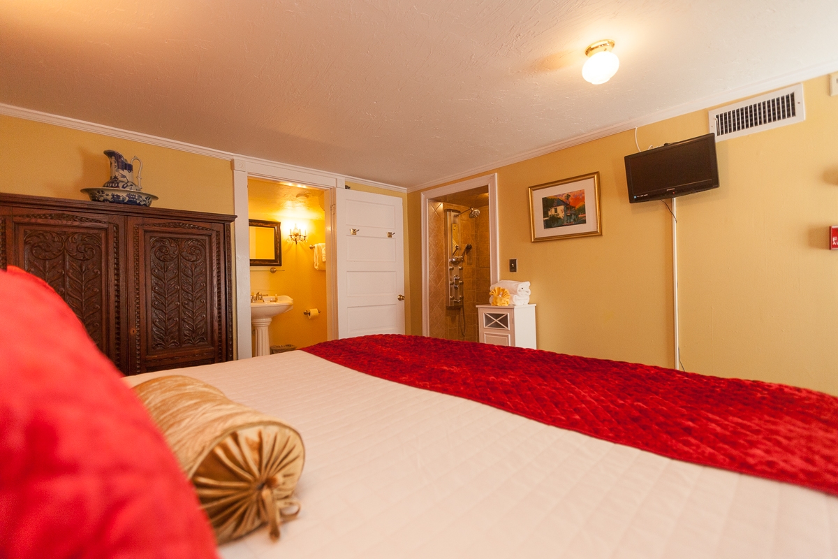 View from plush, sumptuous king bed with white sheets, luxurious red spread and decorative pillows.  Flat-screen TV mounted on wall and doorway leading to private bath.