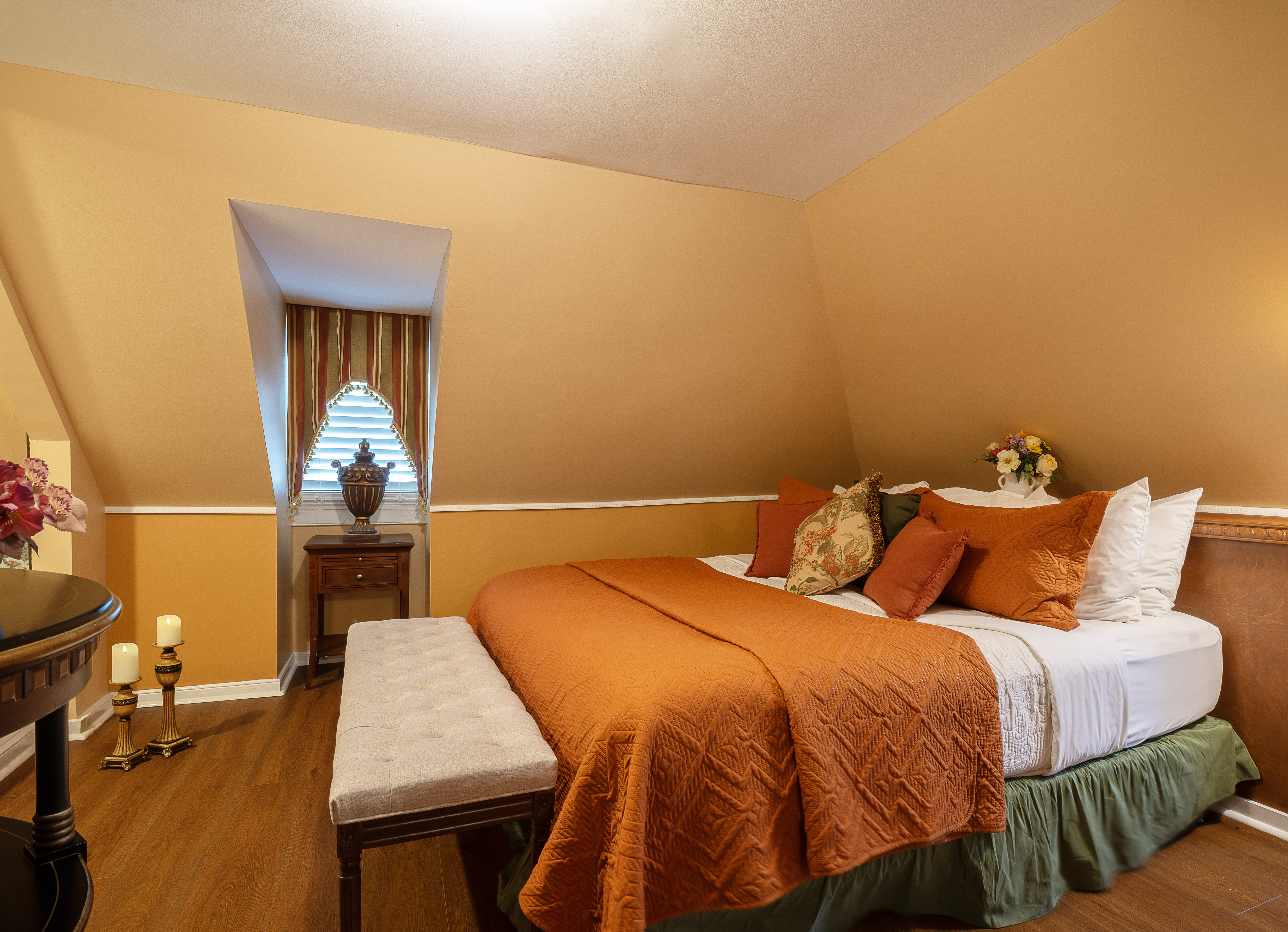 A warm and welcoming third floor bedroom with cheerful walls, finished wood floors, a king bed, and a window seat with plump pillows.