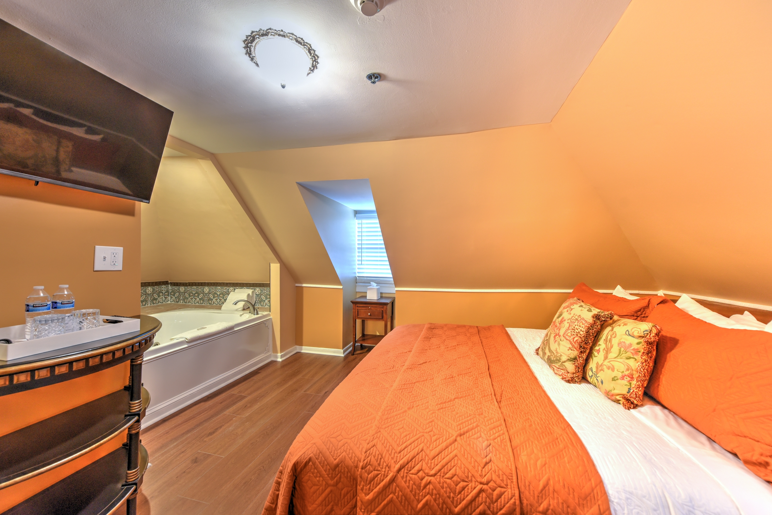 A warm and welcoming third floor bedroom with cheerful walls, finished wood floors, a king bed, and a window seat with plump pillows.