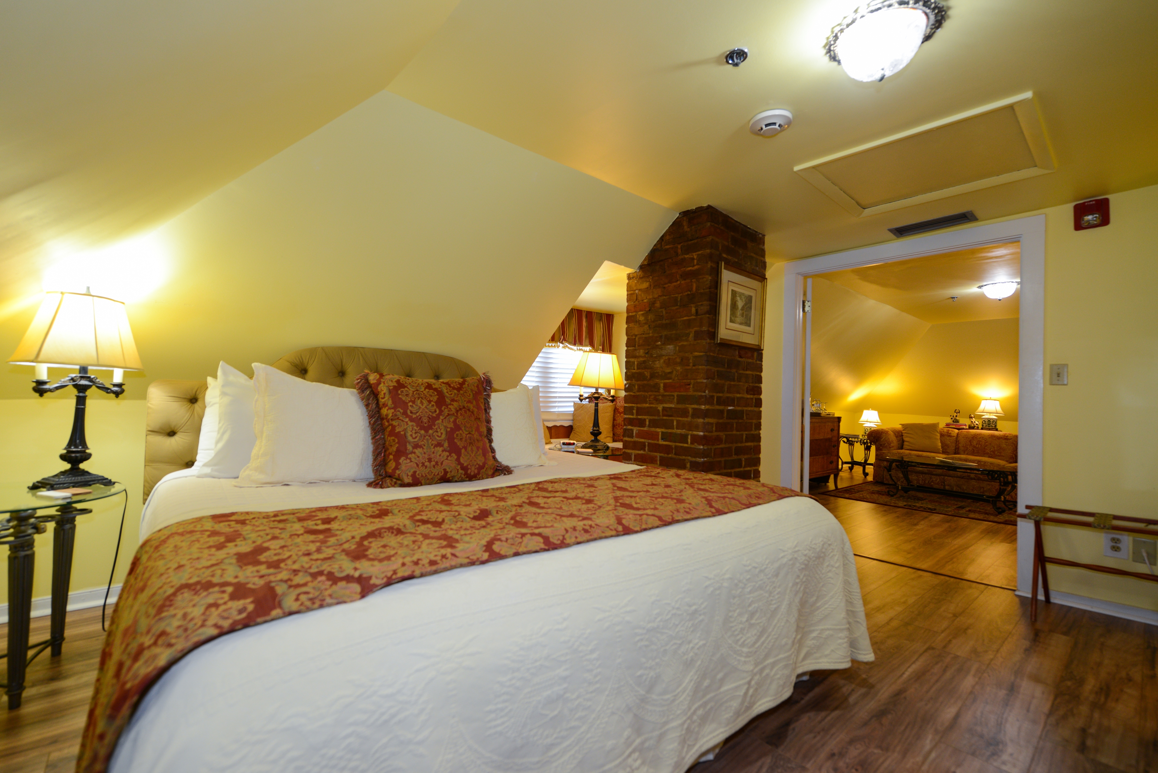 Third-floor bedroom with yellow walls, white trim, and finished wood floors.  The king-size bed has an upholstered tufted headboard with decorative pillows.  The ceiling slopes down just above the headboard, and there is a table lamp on the nightstand.