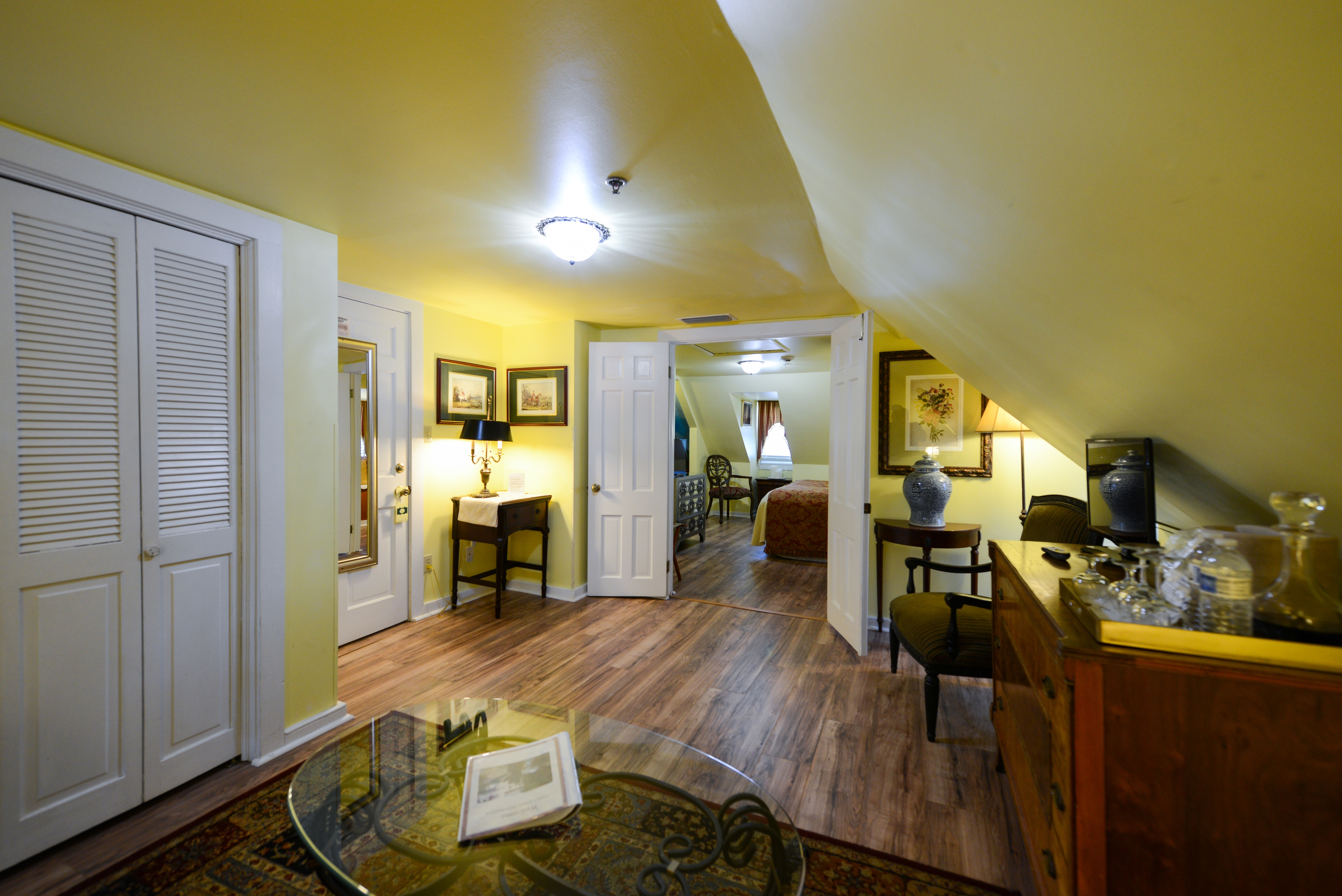 French doors stand open to the bedroom from an inviting sitting room with yellow walls and white trim, finished wood floors, sloped ceilings on one side, and lovely antique furniture.