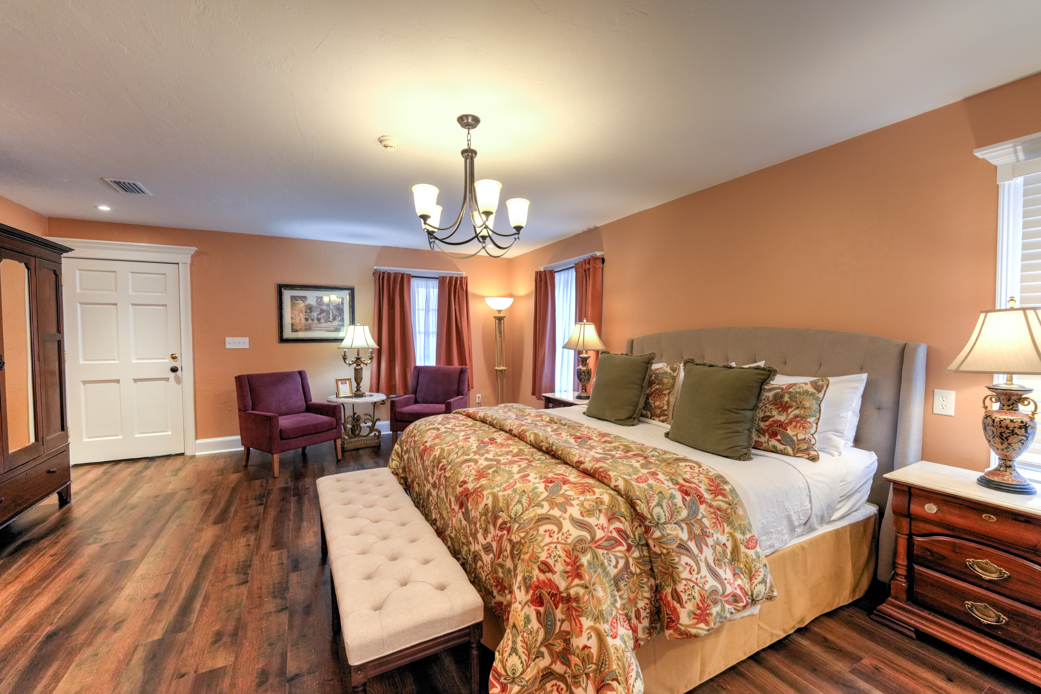 Elegant sun-filled bedroom features a king bed, coordinating night stand with lamp, upholstered bench at foot of bed, and three sunny windows.