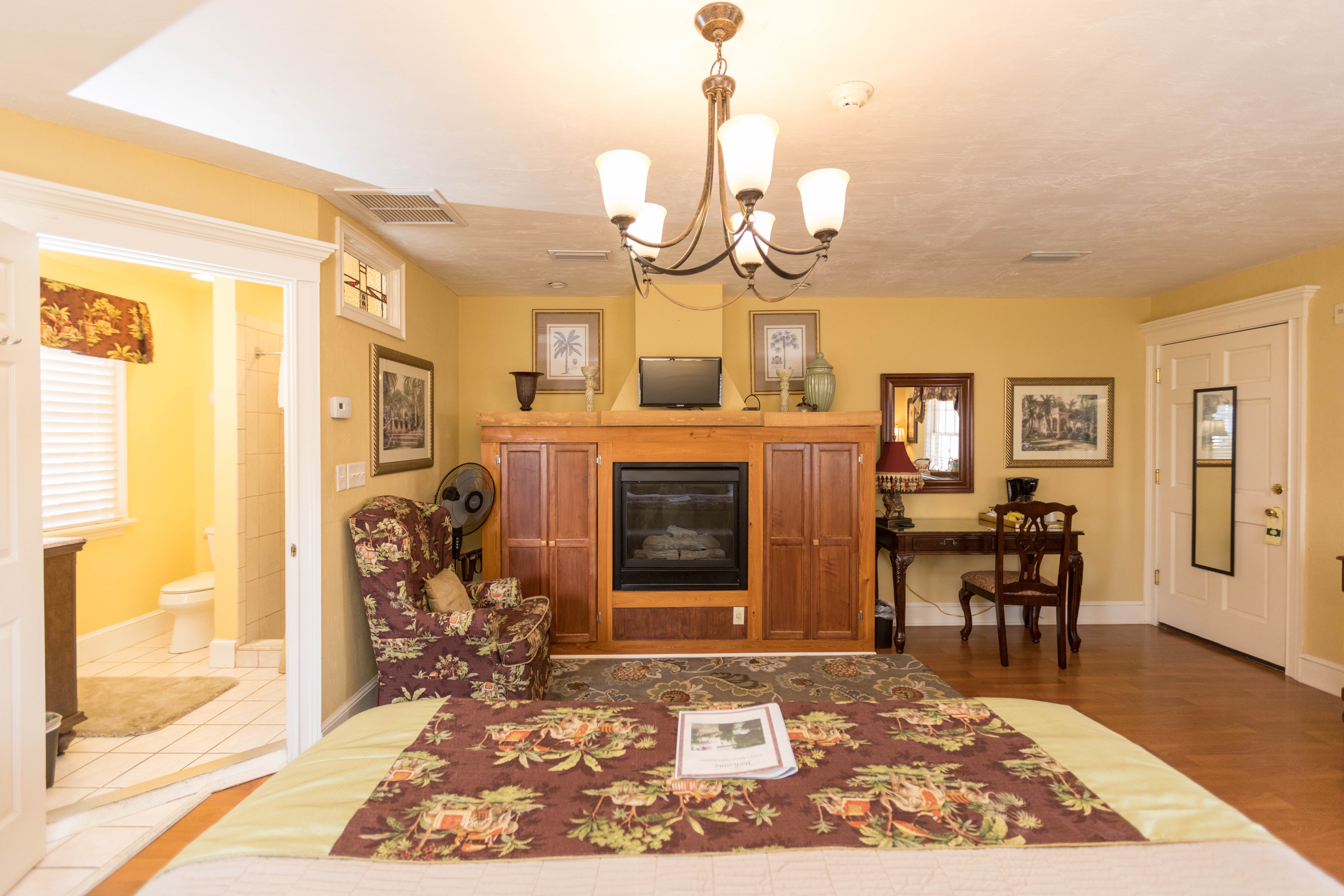 On the wall opposite the bed there is a large wooden cabinet that surrounds the fireplace and also holds a flat-screen TV on top. A floral upholstered chair near the fireplace matches the floral blanket on the bed. To the right of the fireplace is a writing desk with chair.