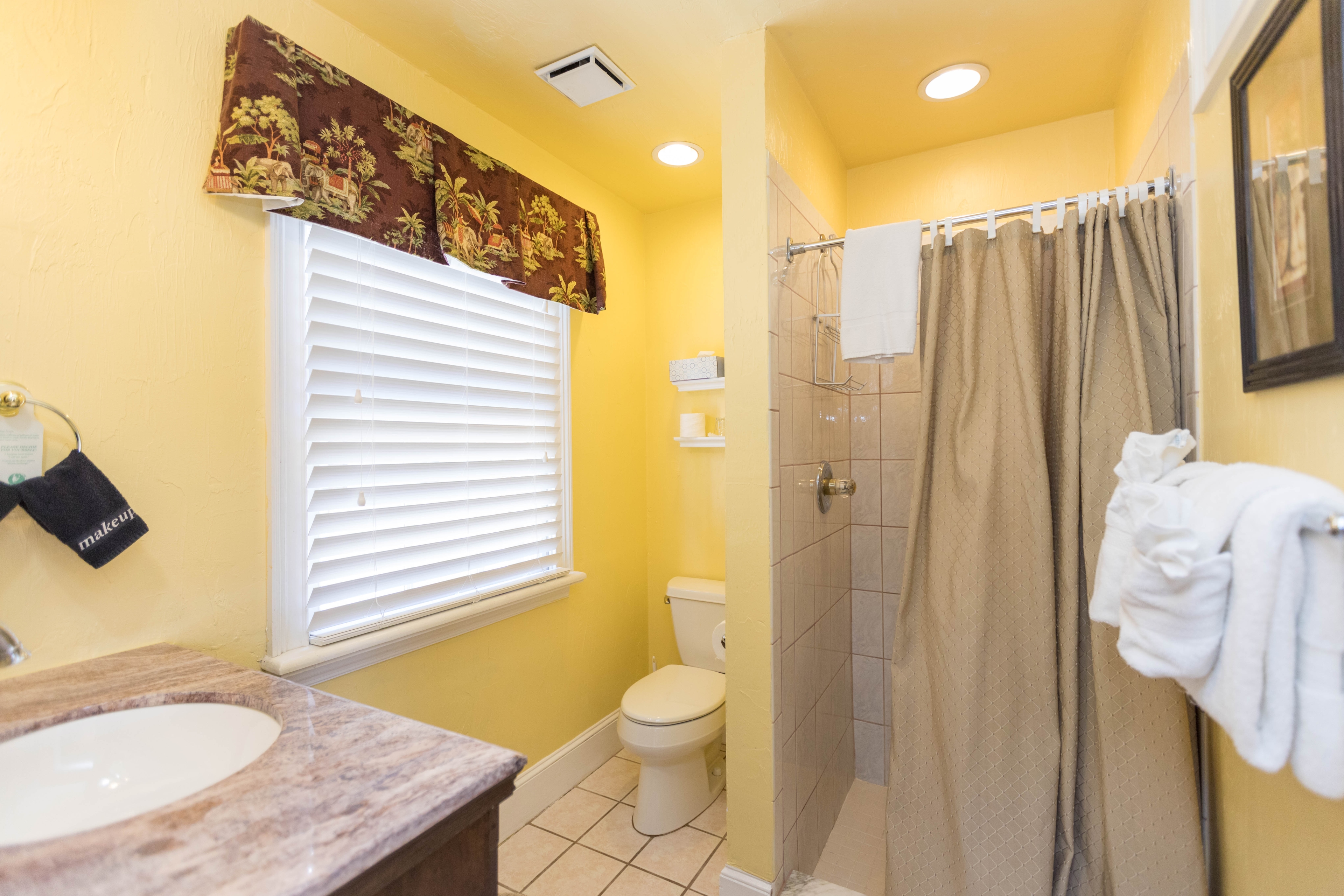 Bright, clean bathroom with walk-in shower, toilet, sink with vanity, and window with blinds and valance.