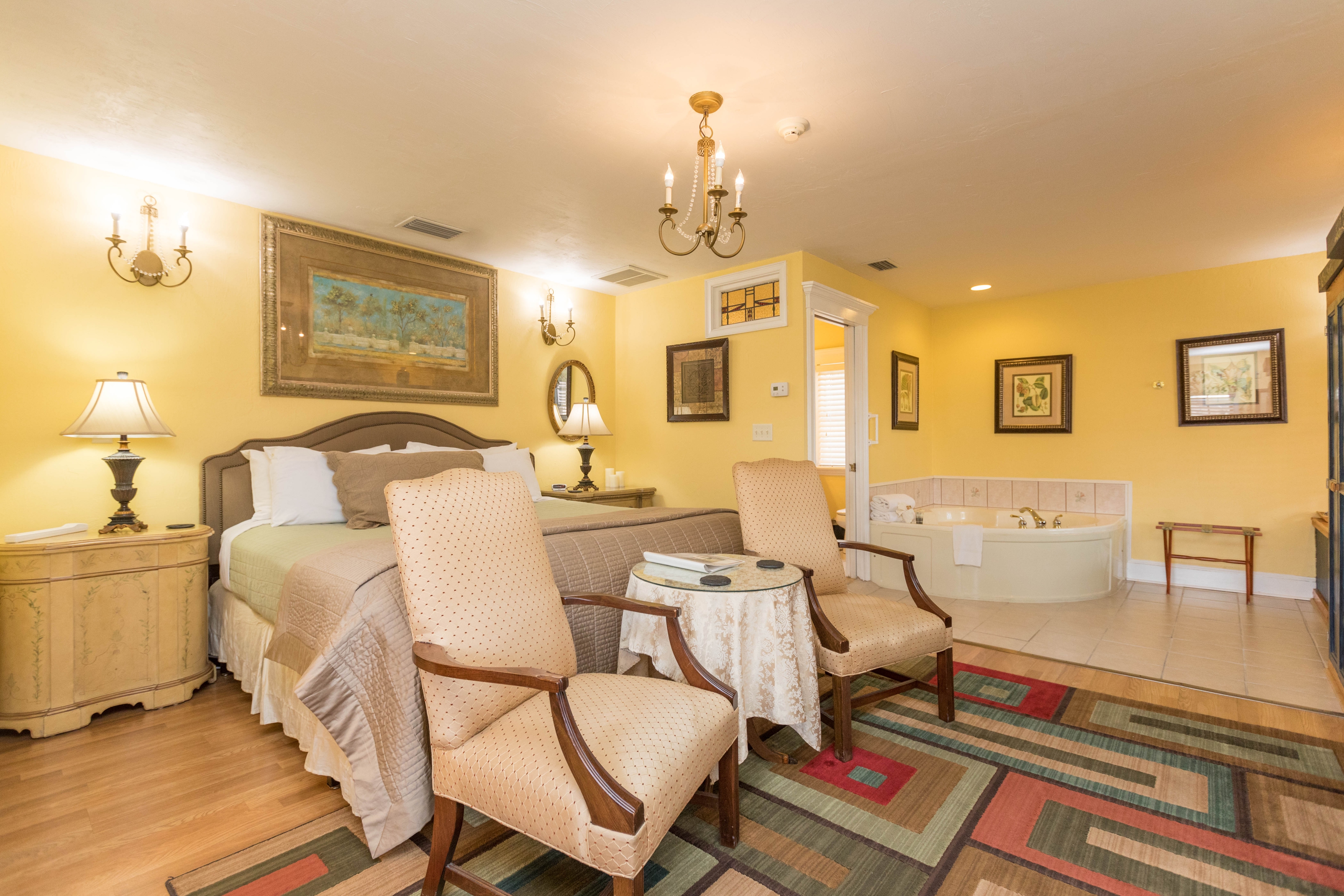 Spacious and elegant bedroom with king bed, ivory colored nightstands with matching lamps, upholstered armchairs with ivory fabric at foot of bed, area rug with bold rectangle pattern, and in the far corner a whirlpool tub.