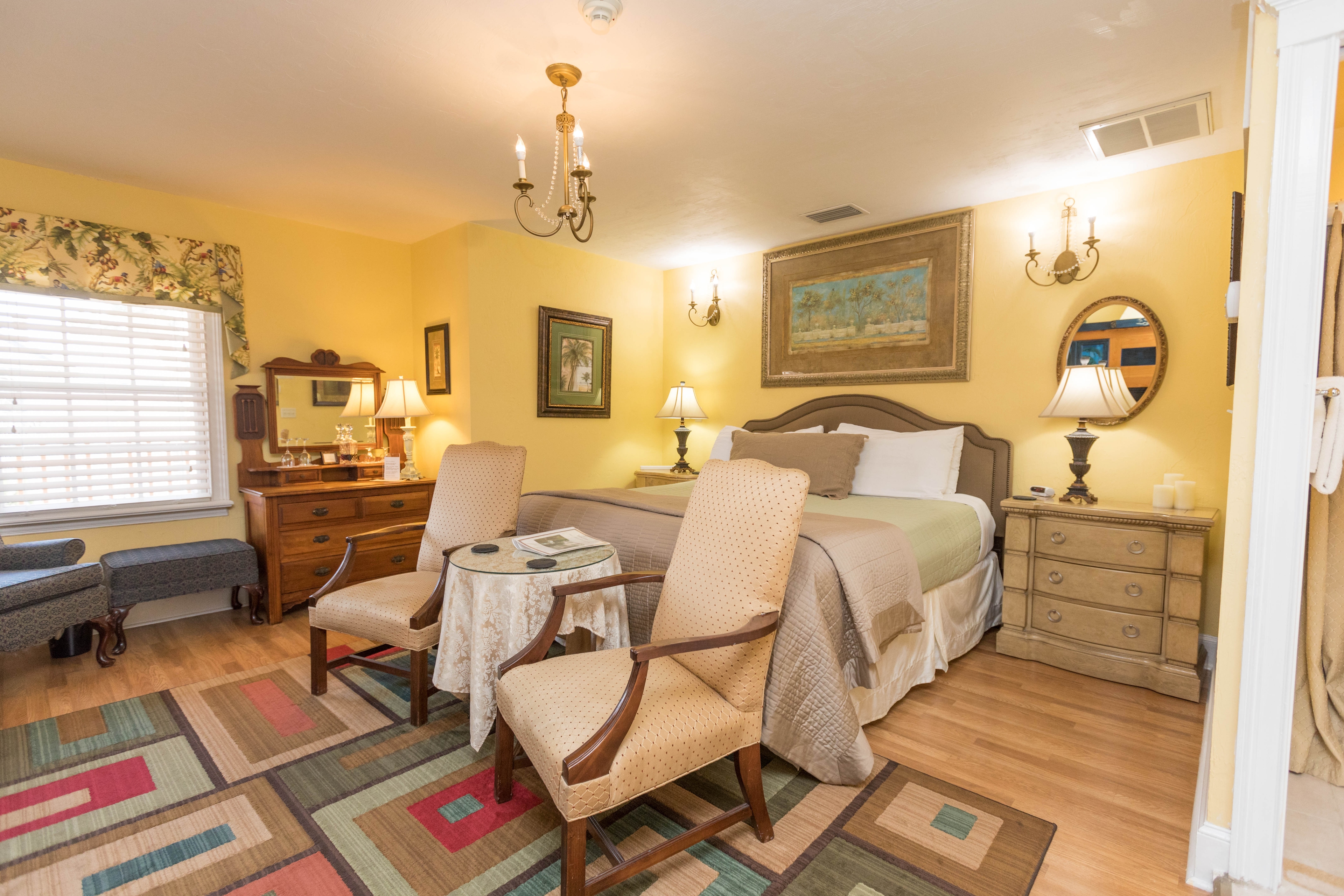 An eclectically decorated room with king bed, matching nightstands with lamps, finished wood floors, two upholstered chairs and an area rug with a bold nested rectangle pattern.