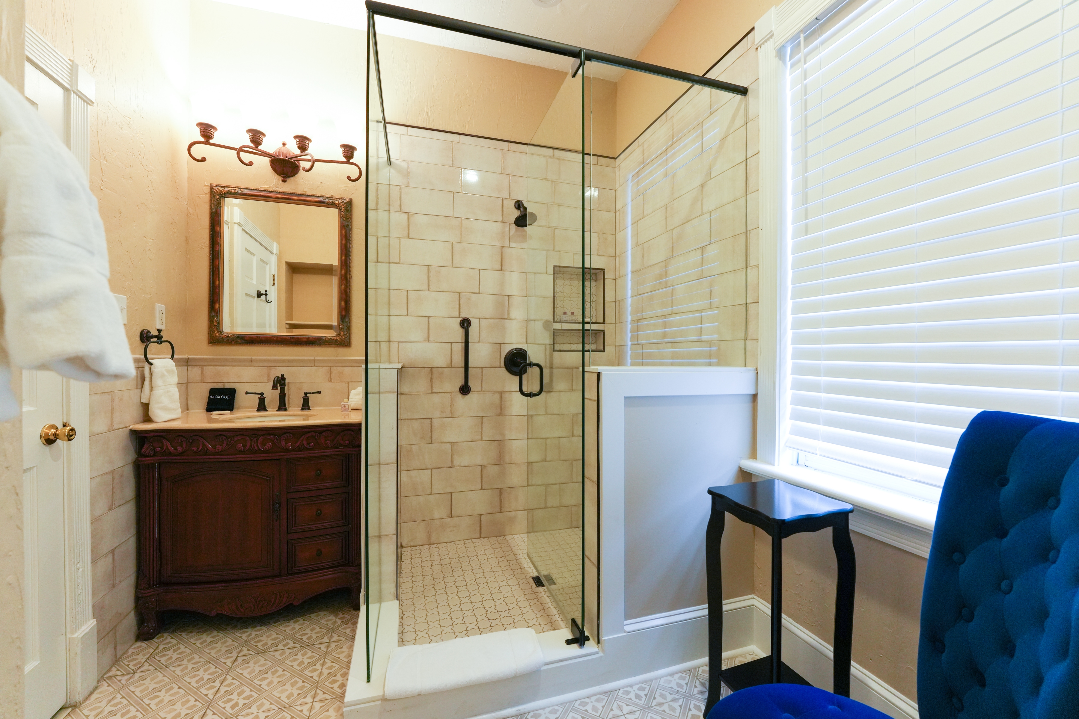 Bright and nicely decorated bathroom with antique vanity, spacious walk-in-shower and high back luxury chair with side table