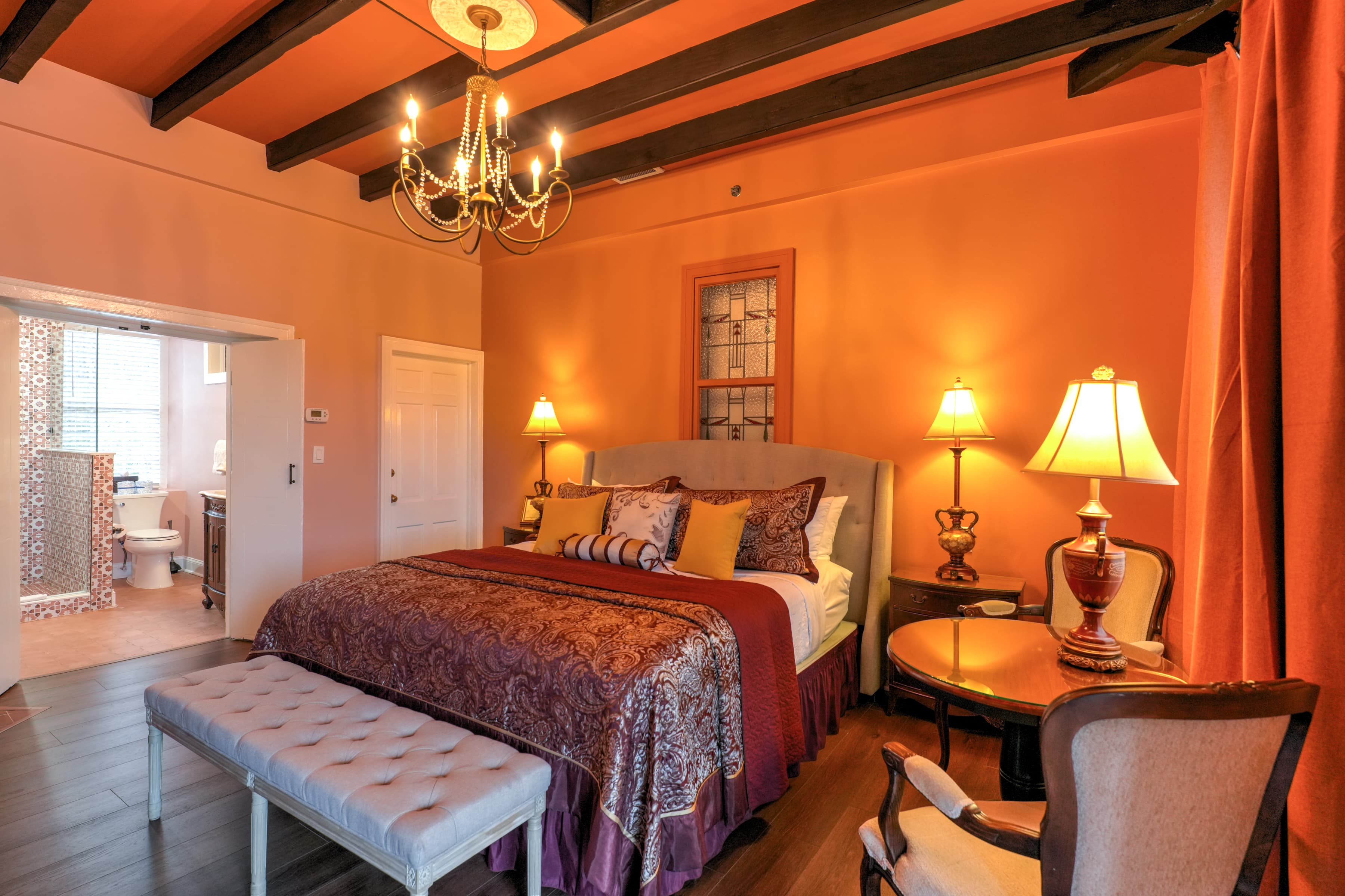 Elegant bedroom with king bed topped with decorative pillows, white sheets, and a floral spread.  The spacious room has a high, beam ceiling and is well appointed with antique furniture and artwork.