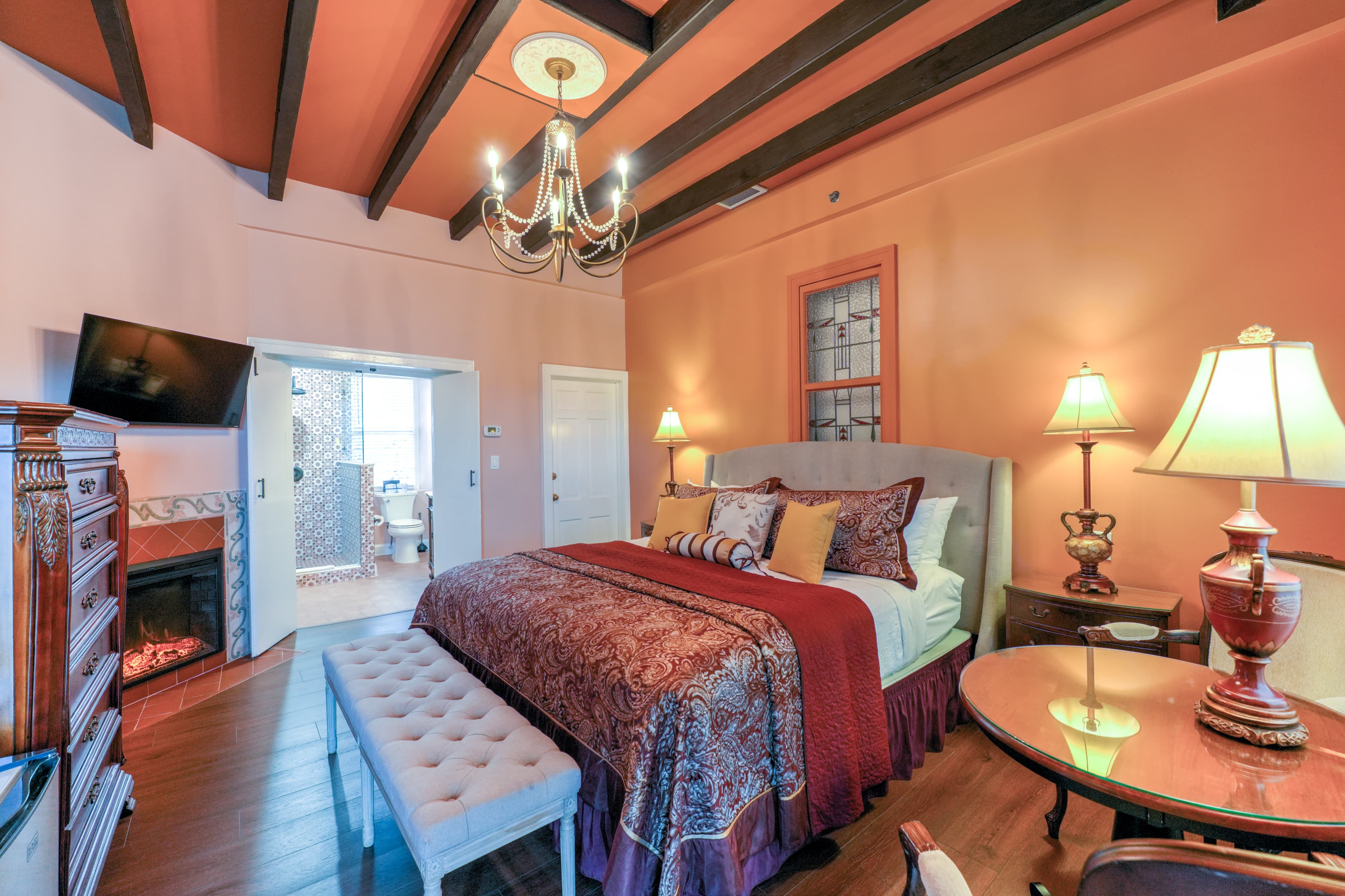 Bedroom with king bed sumptuously appointed with decorative pillows and a floral spread, opposite the bed is a dresser flanked by two antique armchairs and topped with an elegant table lamp