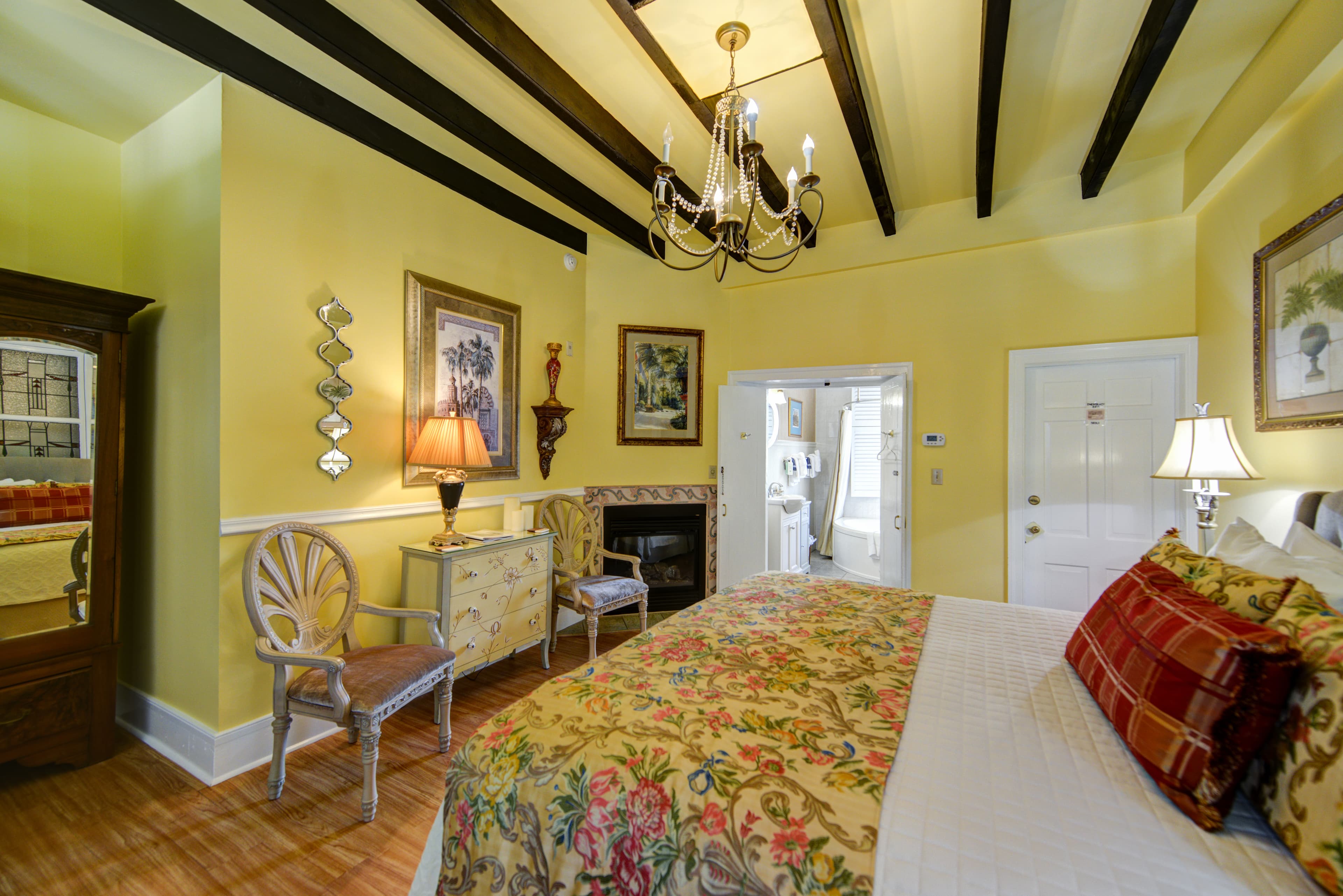 Bedroom with king bed sumptuously appointed with decorative pillows and a floral spread, opposite the bed is a dresser flanked by two antique armchairs and topped with an elegant table lamp