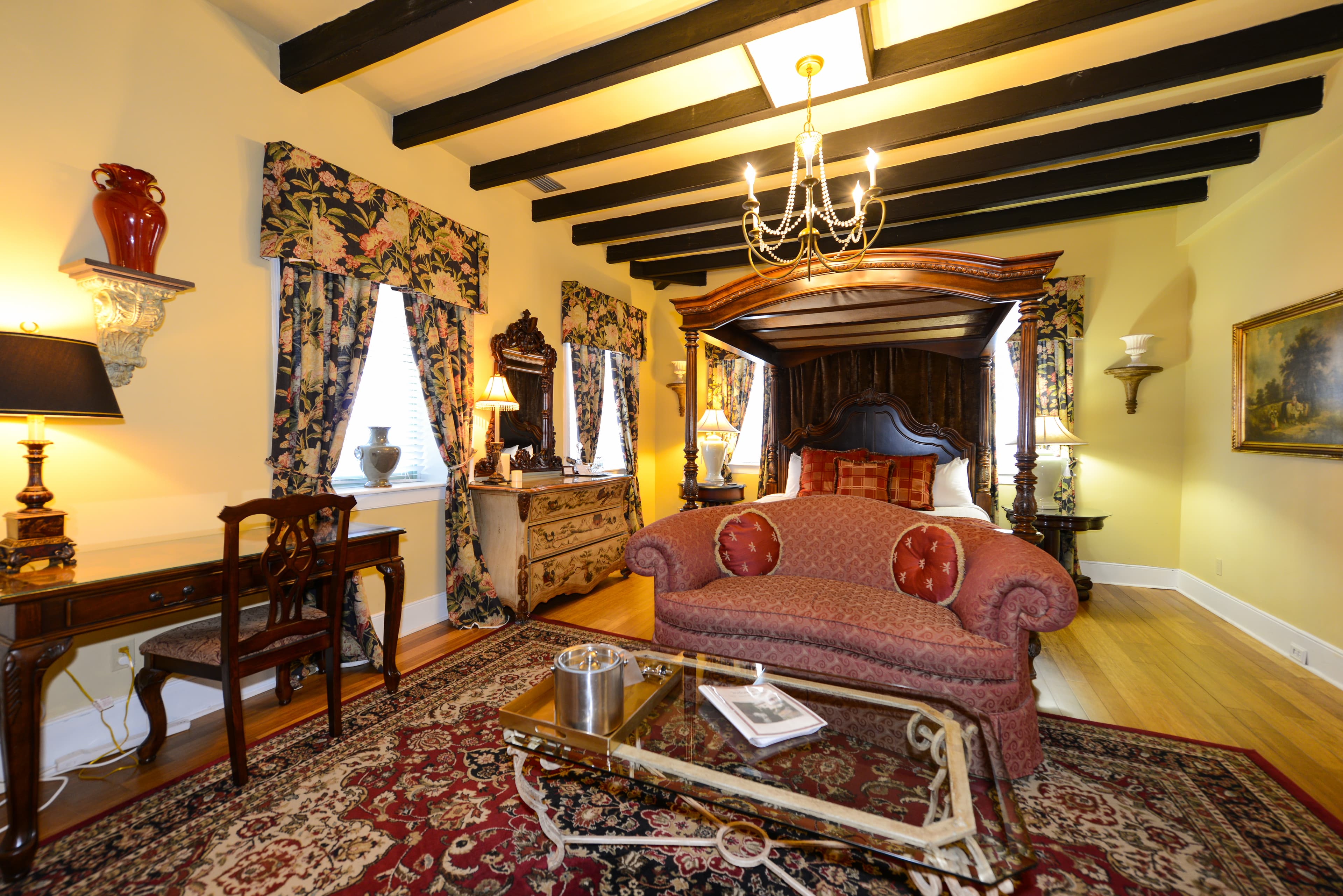 Canopy, four-poster king bed with loveseat at foot of bed, large windows with floral drapes, and writing desk with chair