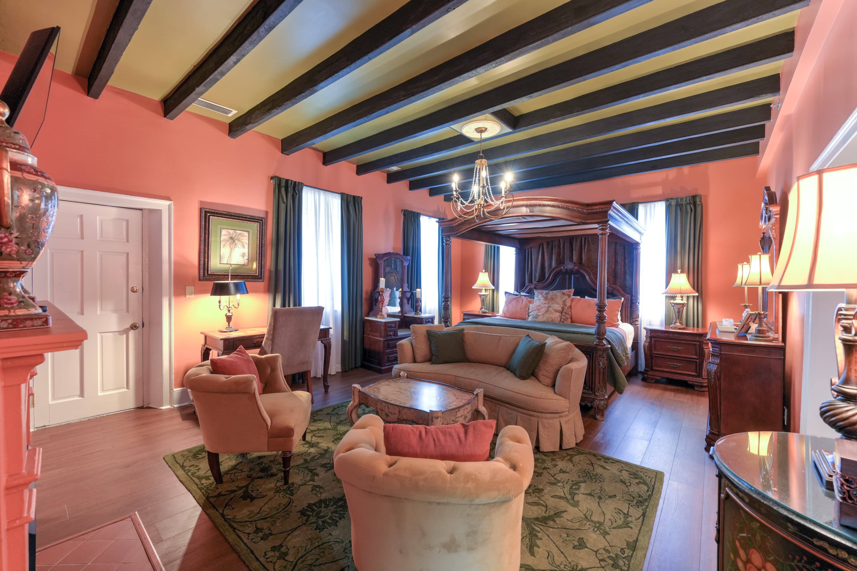 Romantic, luxurious guestroom with canopy four-poster king bed, beam ceiling, and sitting area with oriental area rug and upholstered loveseat and two chairs.  A doorway leads to the private balcony with outdoor sitting area.