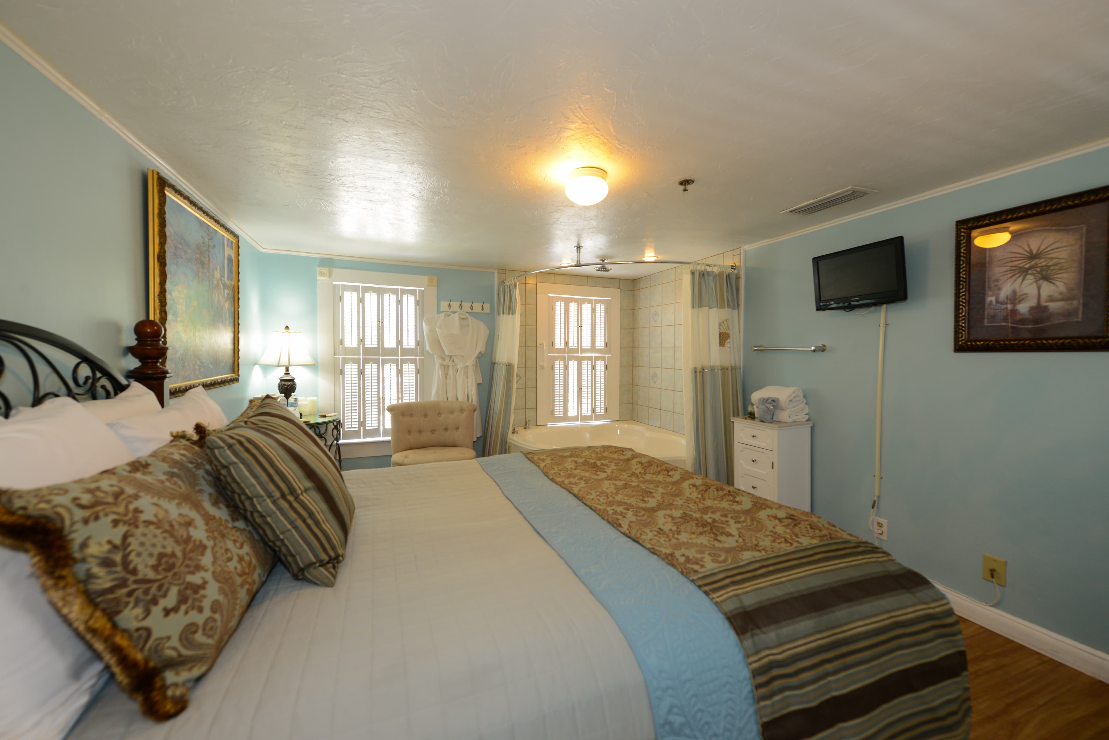 Bedroom with queen bed comfortably topped with blankets and decorative pillows. A flat-screen TV is mounted on the opposite wall, and two large windows let in natural light at the far end of the room.