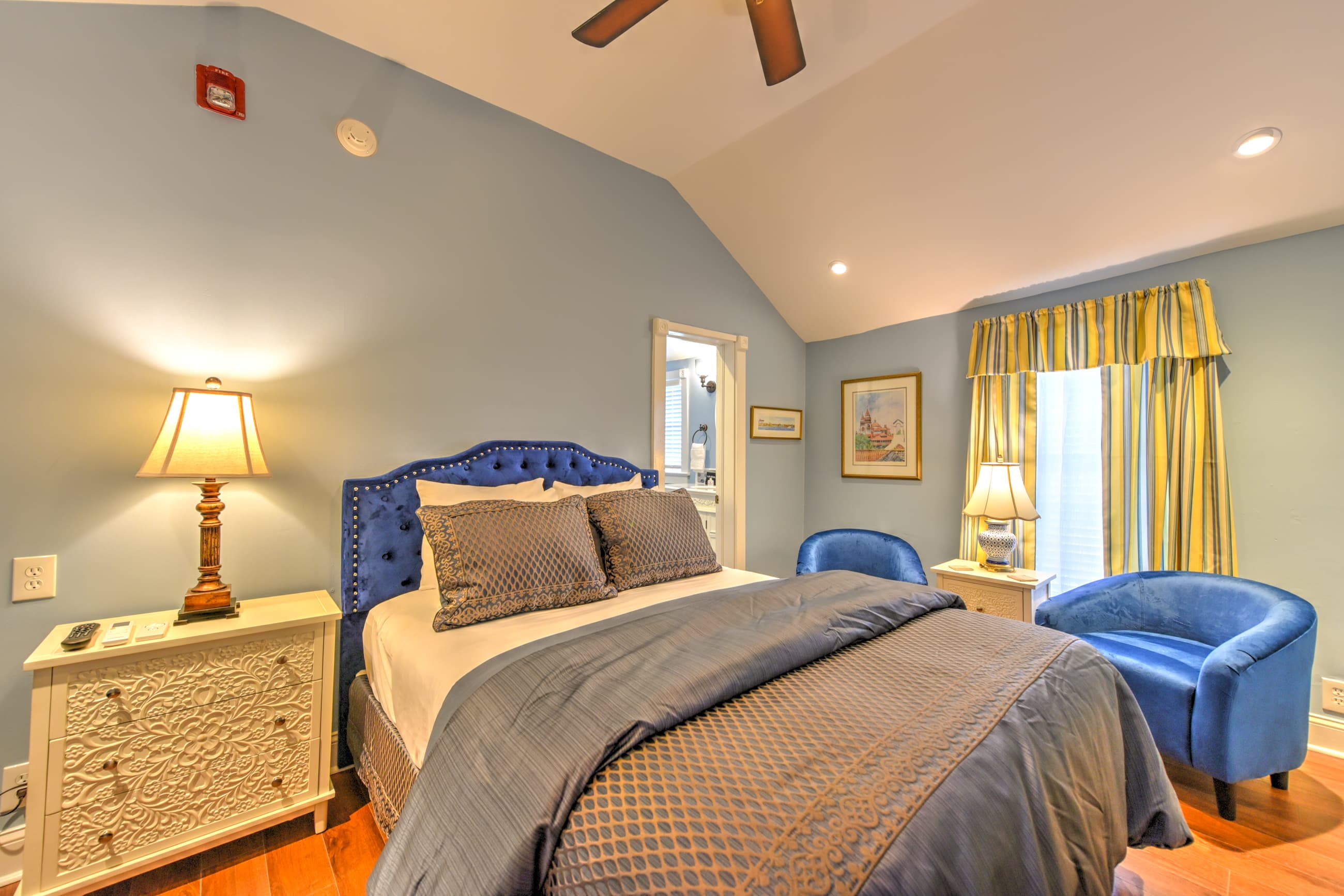 Charming bedroom decorated in blue, yellow, white with a queen bed, sunny window and finished wood floors.