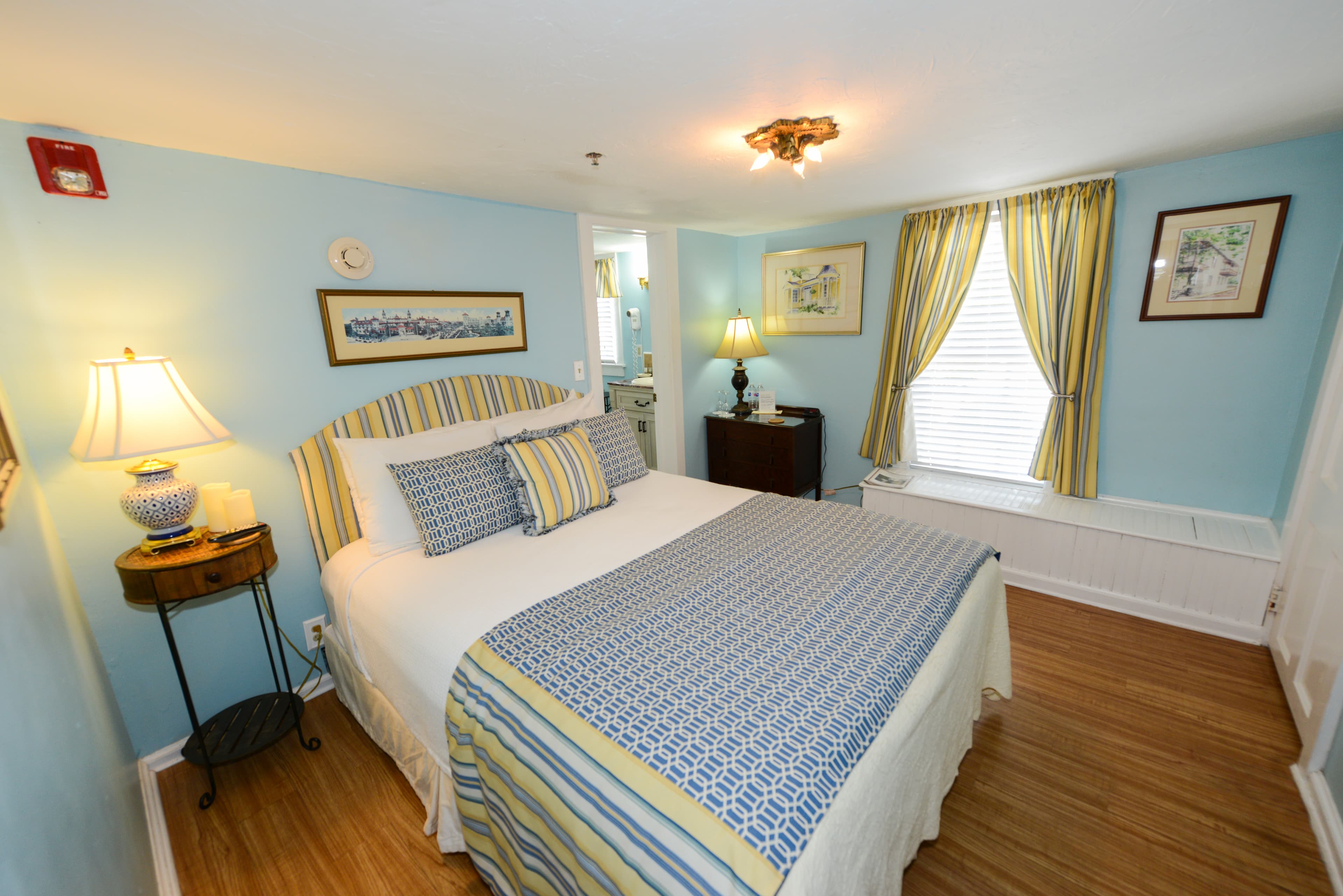 Charming bedroom decorated in light blue, yellow, white with a queen bed, sunny window with built-in window seat, and finished wood floors.