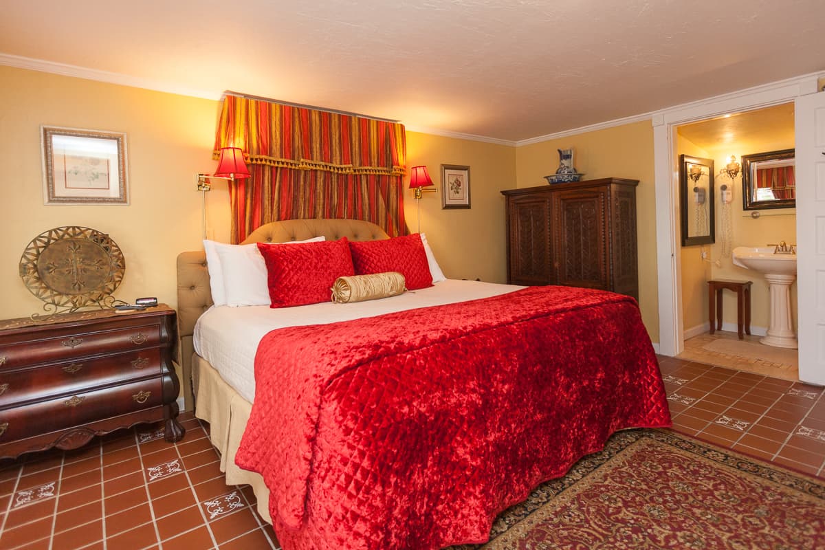 Warm and inviting first-floor bedroom with king bed topped with rich red luxe bedspread, decorative throw pillows and tufted upholstered headboard, dresser, armoire, and doorway to private bath.