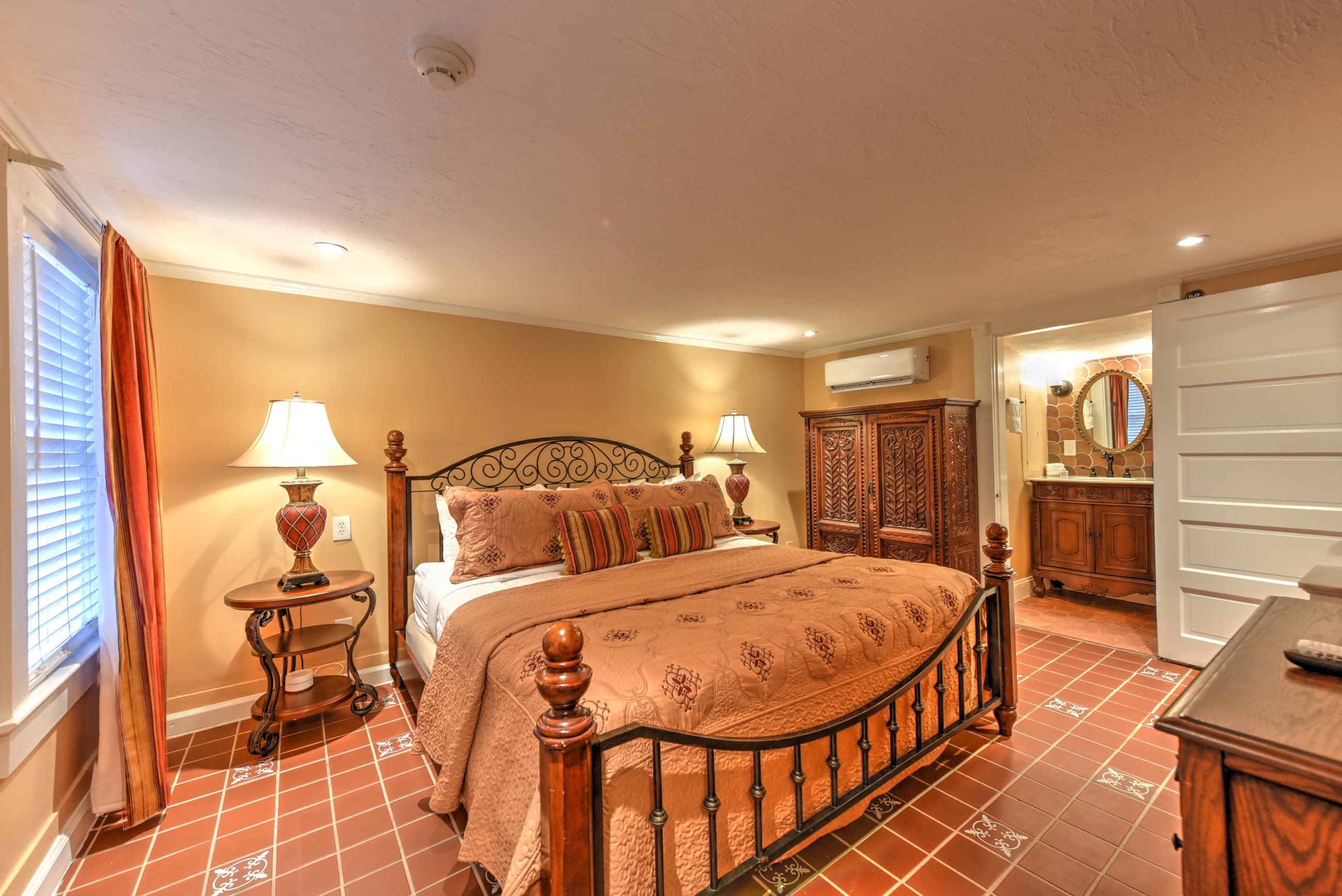 Warm and inviting first-floor bedroom with king bed topped with rich red luxe bedspread, decorative throw pillows, dresser, armoire, and doorway to private bath.