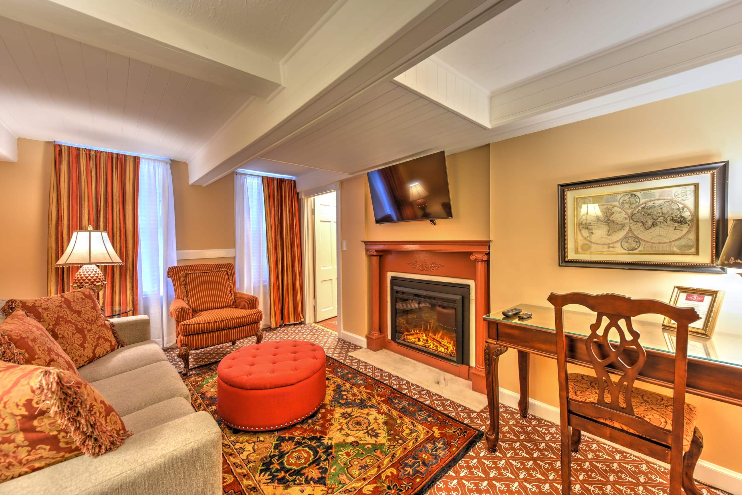 Sitting area decorated in shades of rich red and earth tones and comfortably appointed with sofa, armchairs and ottoman. Other features include two sunny windows with elegant drapes, beam ceiling, and fireplace with painted wood mantel.