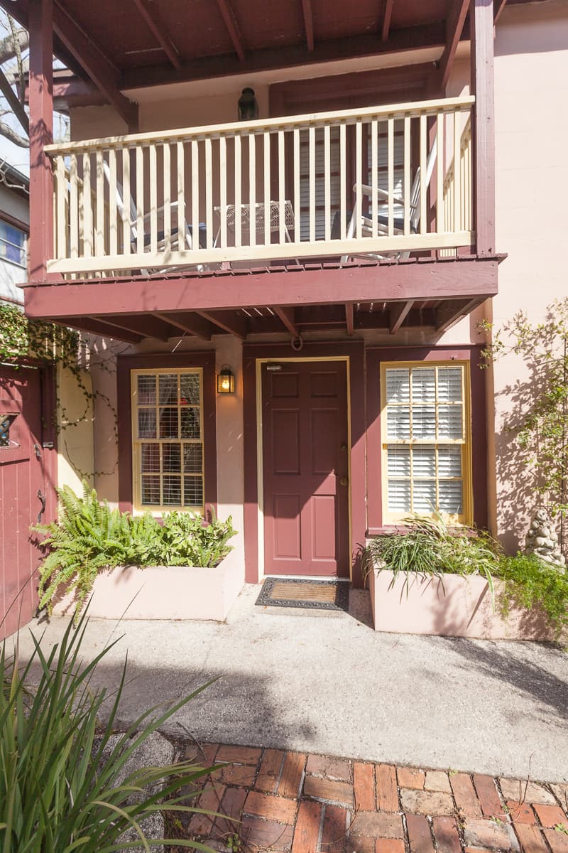 Exterior of inn with doorway leading to guestroom flanked with rectangular planters outside large windows and second-floor private balcony with rocking chairs.