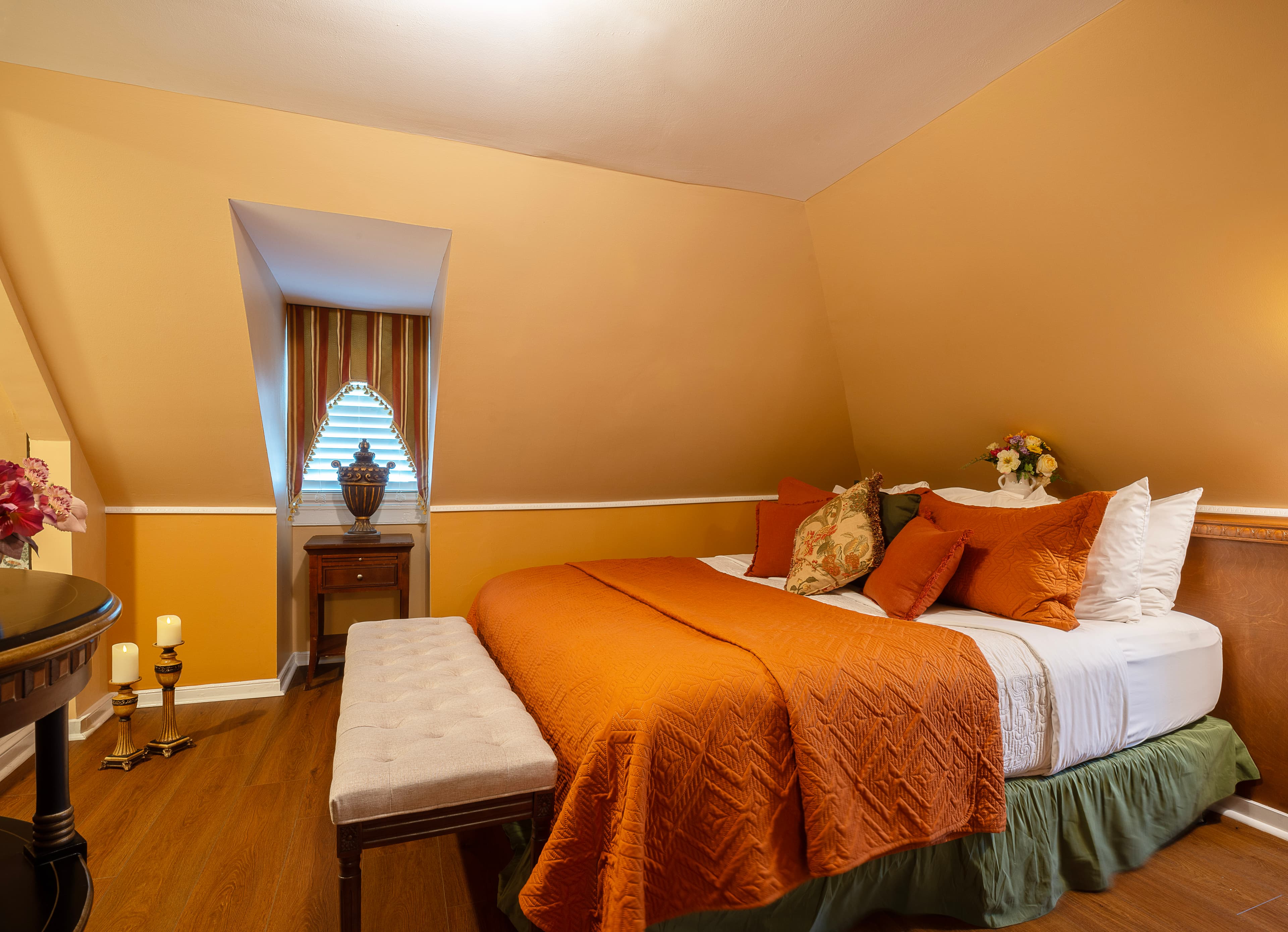 A warm and welcoming third floor bedroom with cheerful walls, finished wood floors, a king bed, and a window seat with plump pillows.