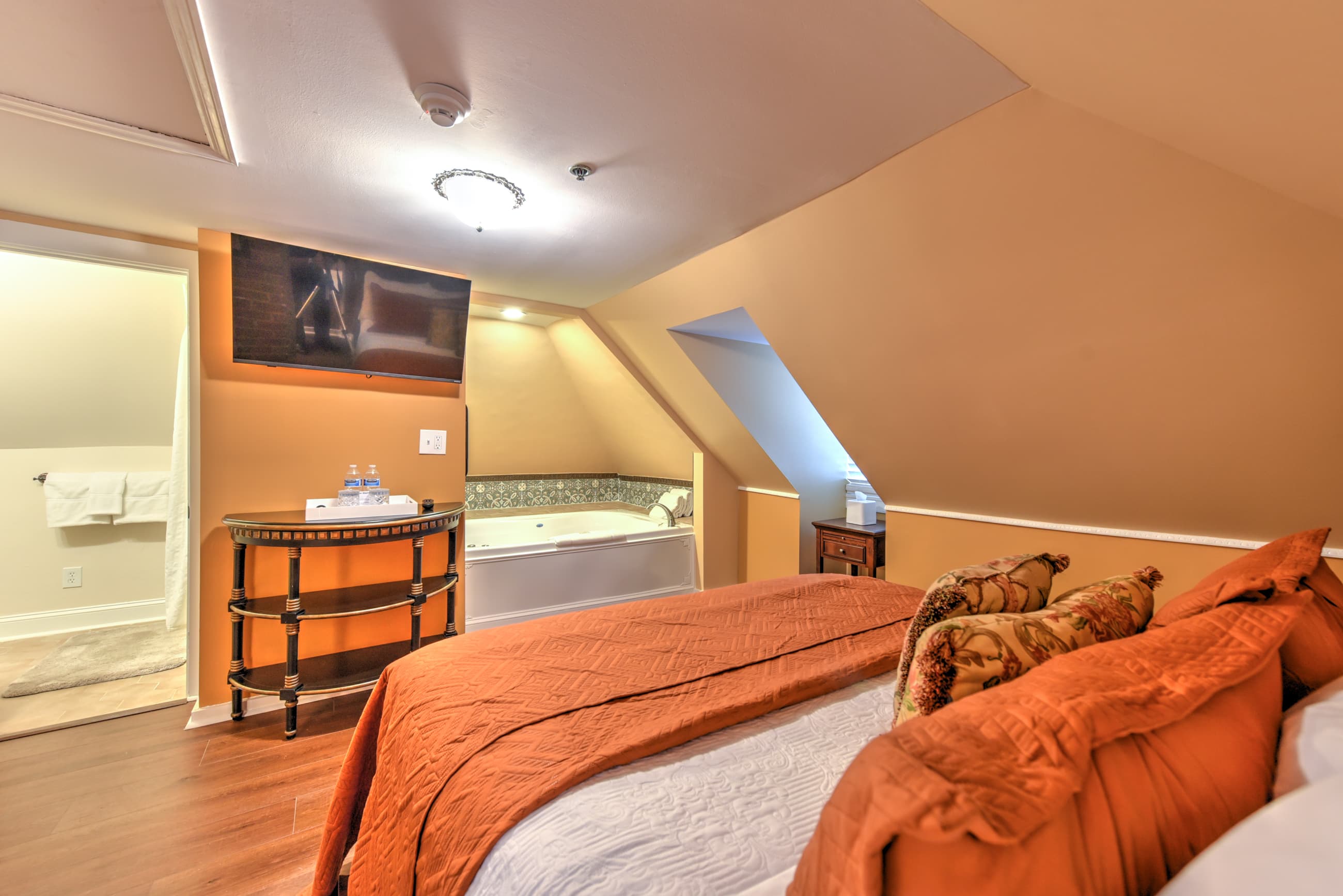 Third-floor bedroom with yellow walls, white trim, and finished wood floors.  The king-size bed with decorative pillows. The ceiling slopes down just above the headboard, and there is a table lamp on the nightstand.