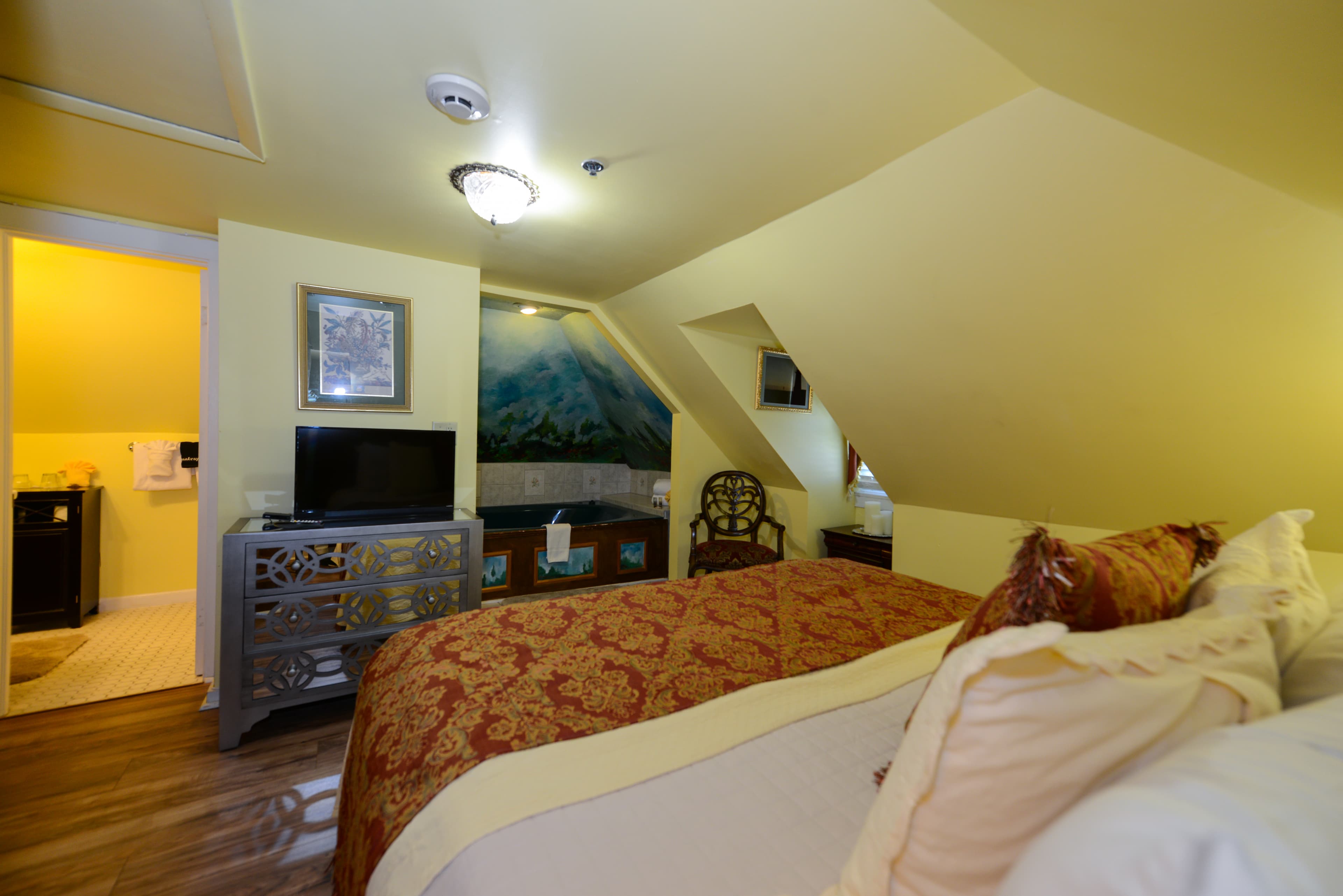 Cozy third-floor bedroom opposite the king bed is a bureau with a flat-screen TV.  To the left of the TV is the bathroom doorway, and recessed in a nook to the right is a whirlpool tub for two.