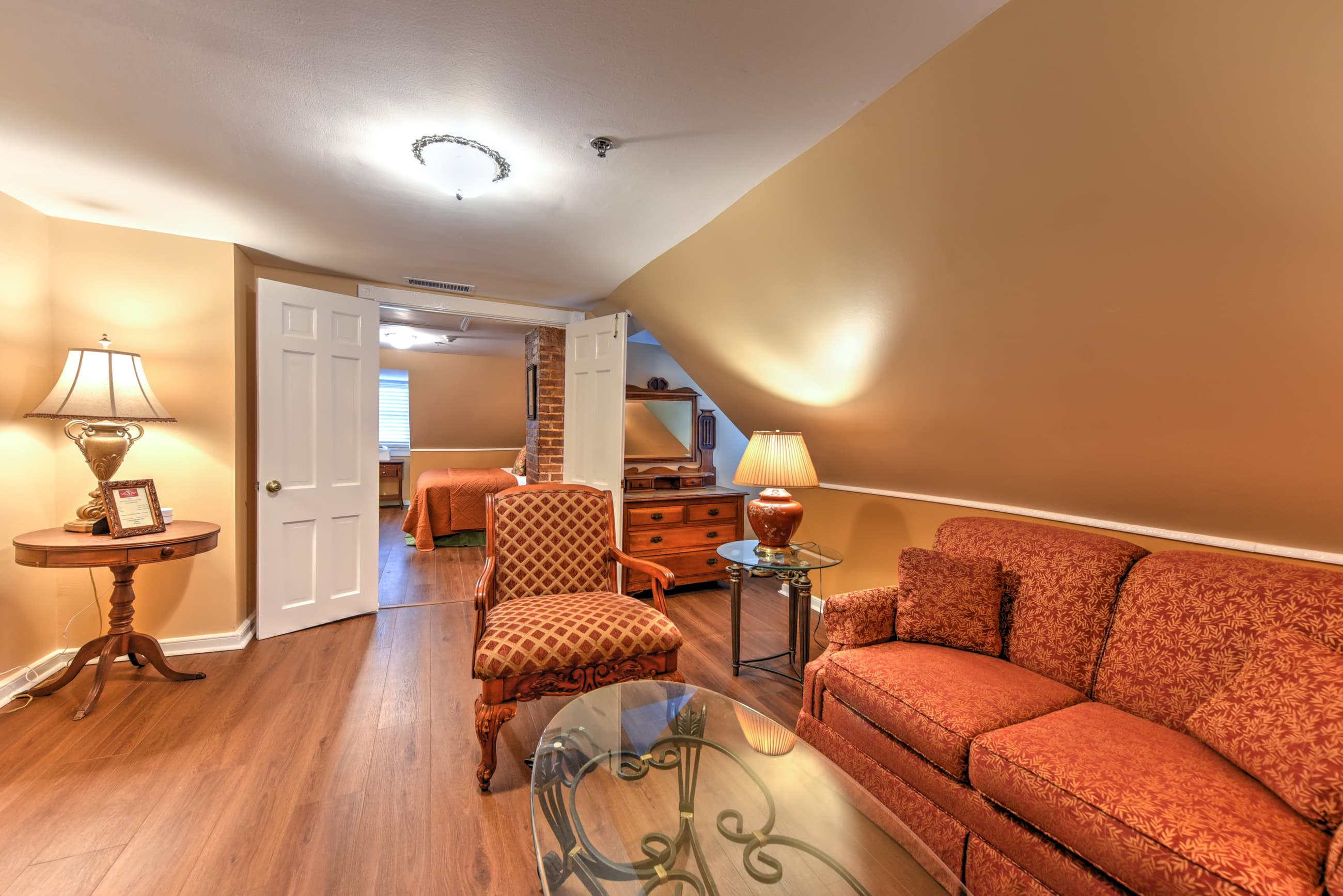 French doors stand open to the bedroom from an inviting sitting room with cheering walls and white trim, finished wood floors, sloped ceilings on one side, and lovely antique furniture.