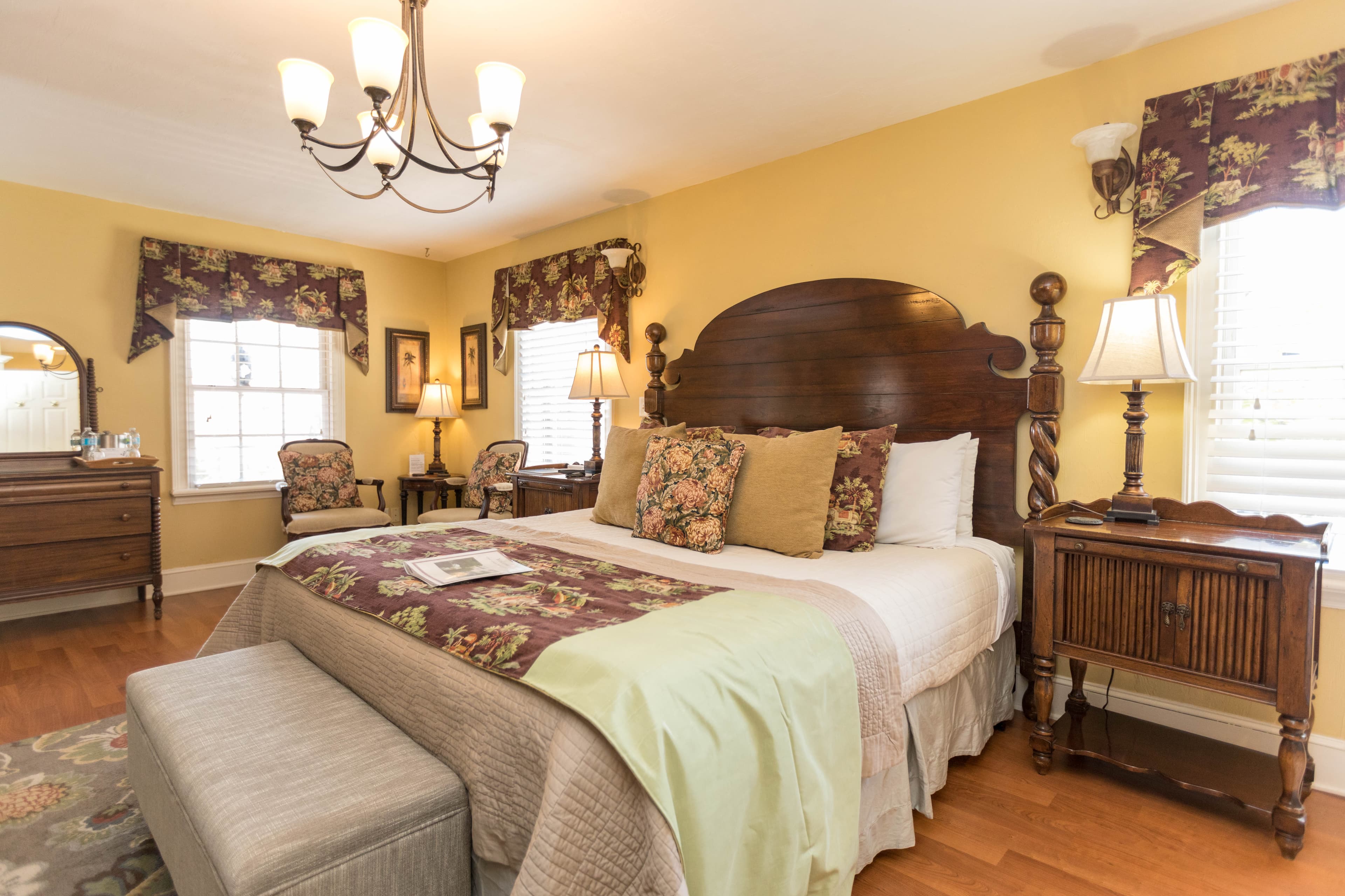 Elegant sun-filled bedroom features a king bed with large wooden antique-style headboard, coordinating night stand with lamp, upholstered bench at foot of bed, and three sunny windows.