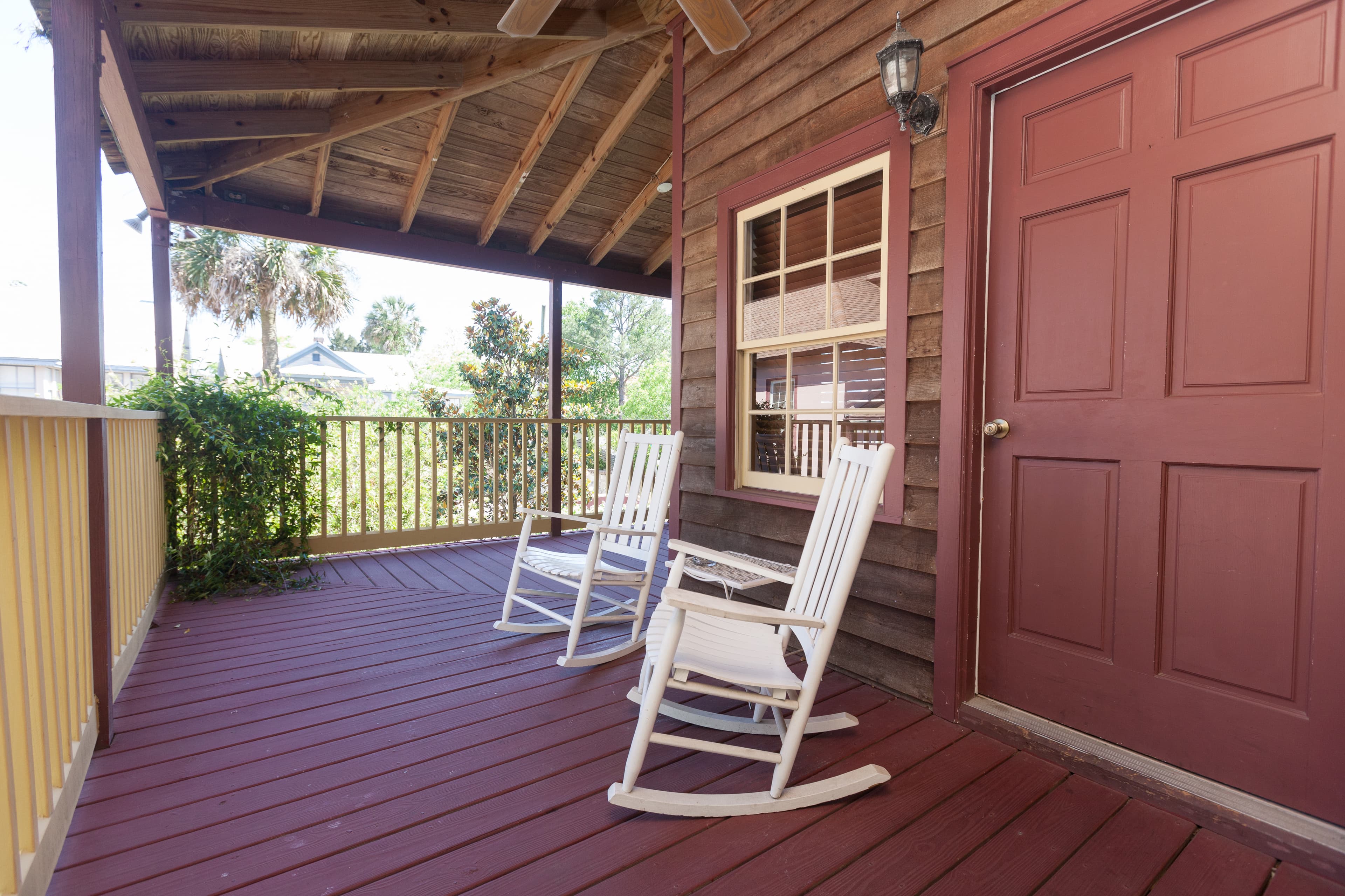 Private section of wrap-around porch painted a deep red with two white rocking chairs and a doorway leading to guestroom.