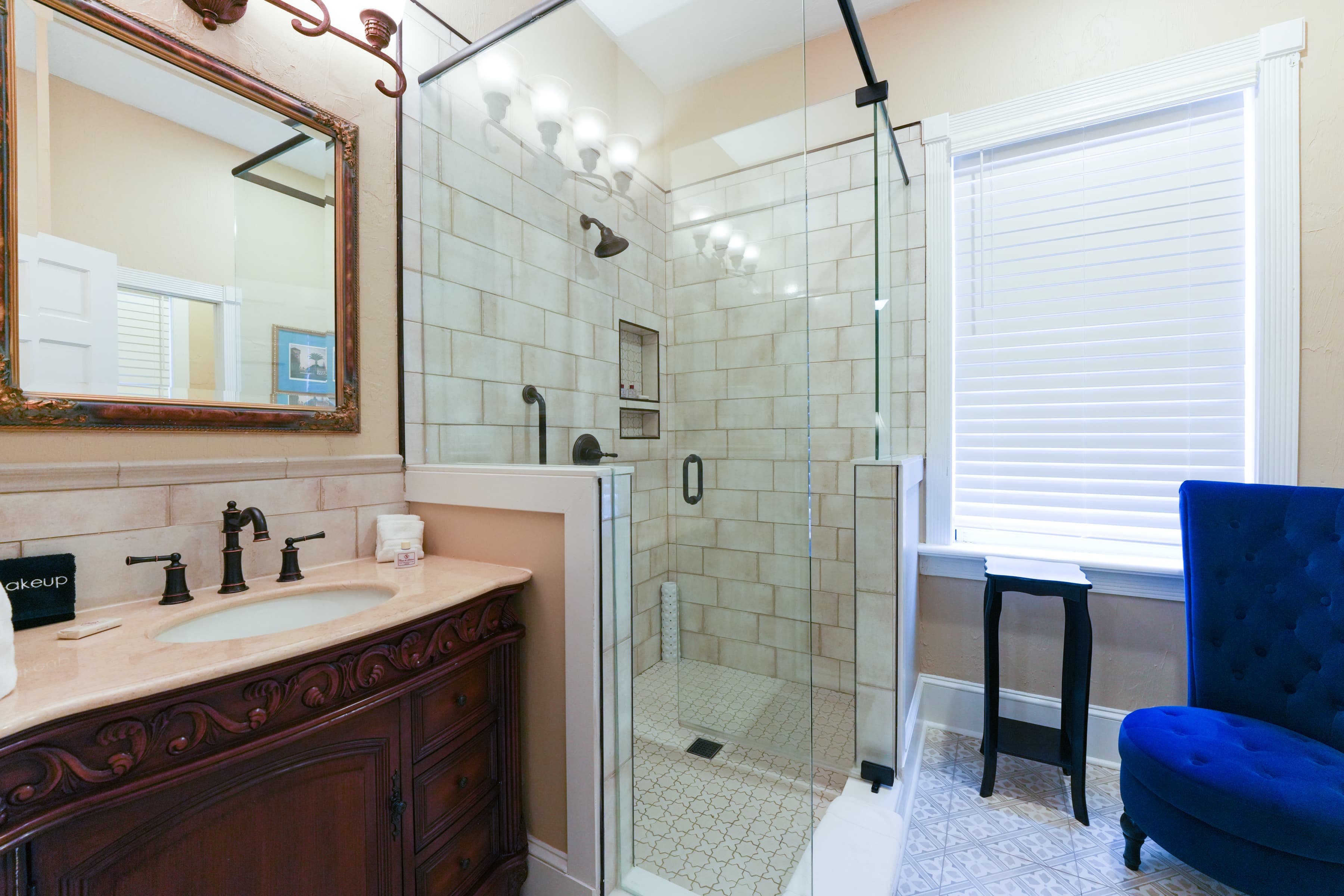 Bright and nicely decorated bathroom with antique vanity, spacious walk-in-shower and high back luxury chair with side table