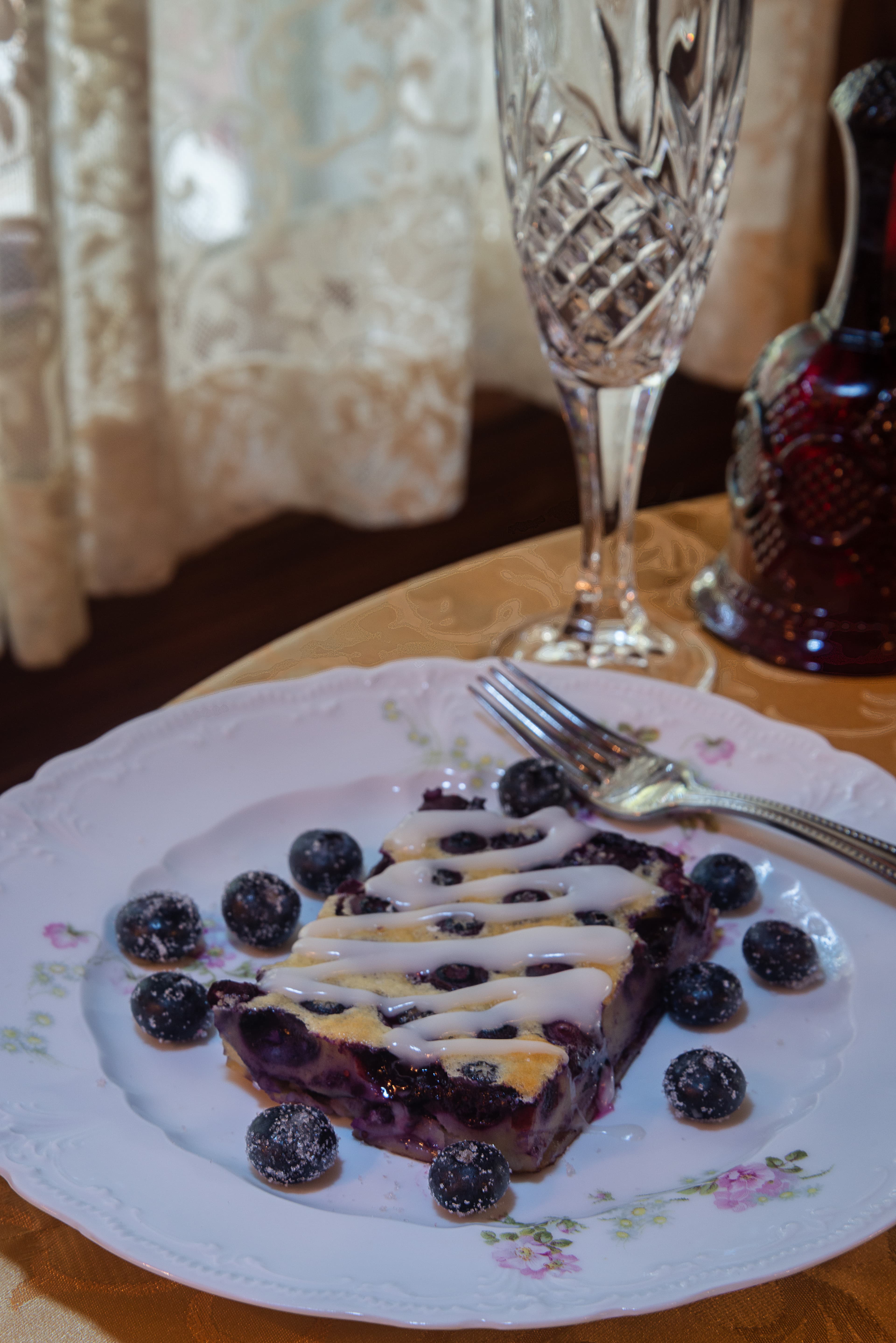 A slice of blueberry dessert drizzled with icing, surrounded by fresh blueberries, on a decorative plate.
