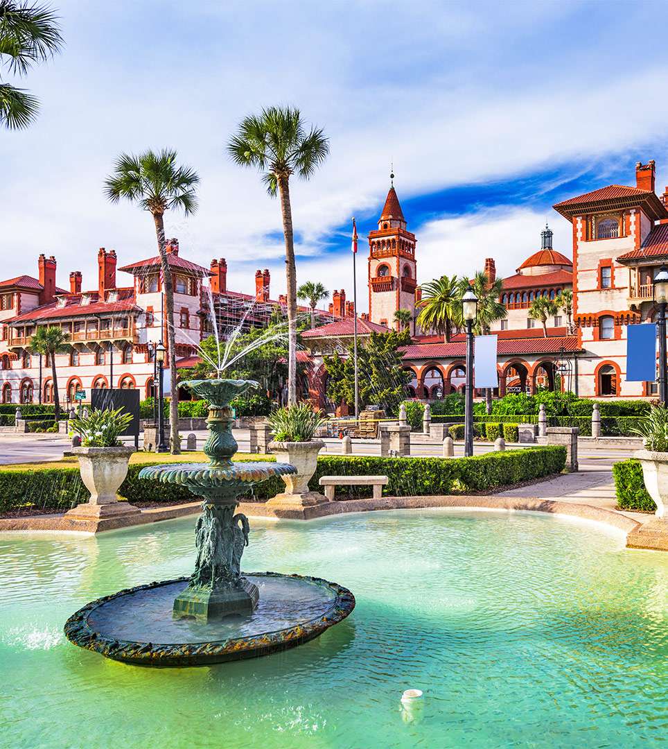 A decorative fountain in a lush courtyard featuring historical buildings and palm trees under a blue sky.