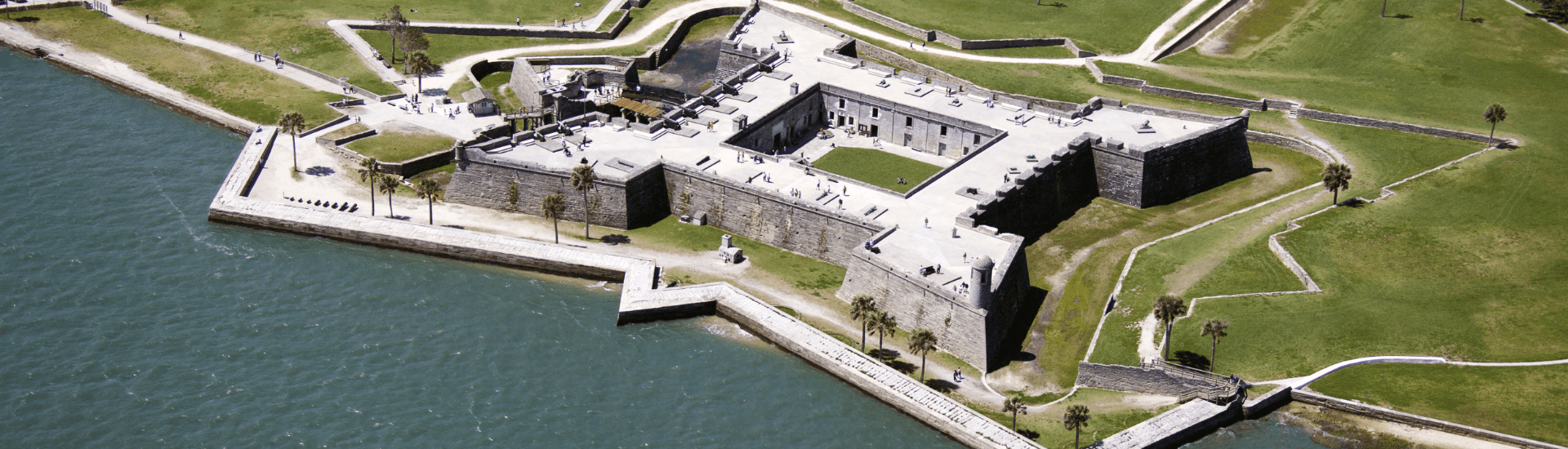 An aerial view of Castillo de San Marcos National Monument in St. Augustine, Florida. The fort is a large, stone structure surrounded by water on three sides. There are walkways, buildings, and trees within the fort.