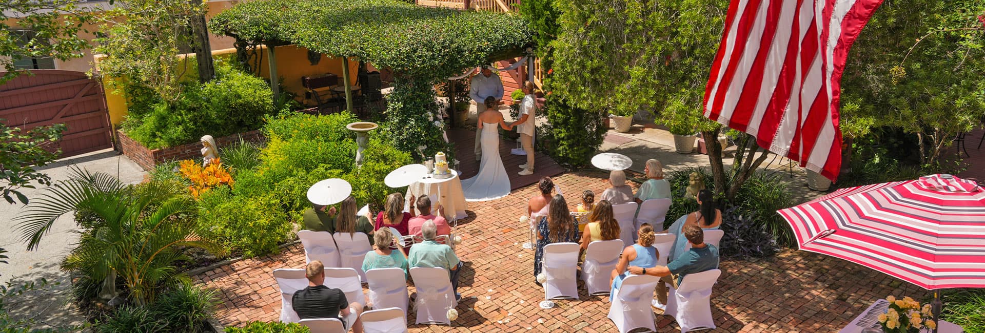 A wedding ceremony taking place outdoors, with guests seated on a brick patio under greenery and an American flag.