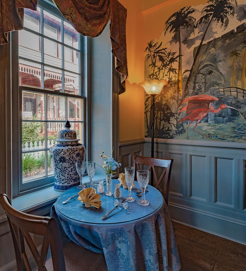 A blue table set for dining with crystal glasses and decorative elements, next to a window featuring a scenic mural and a lamp.