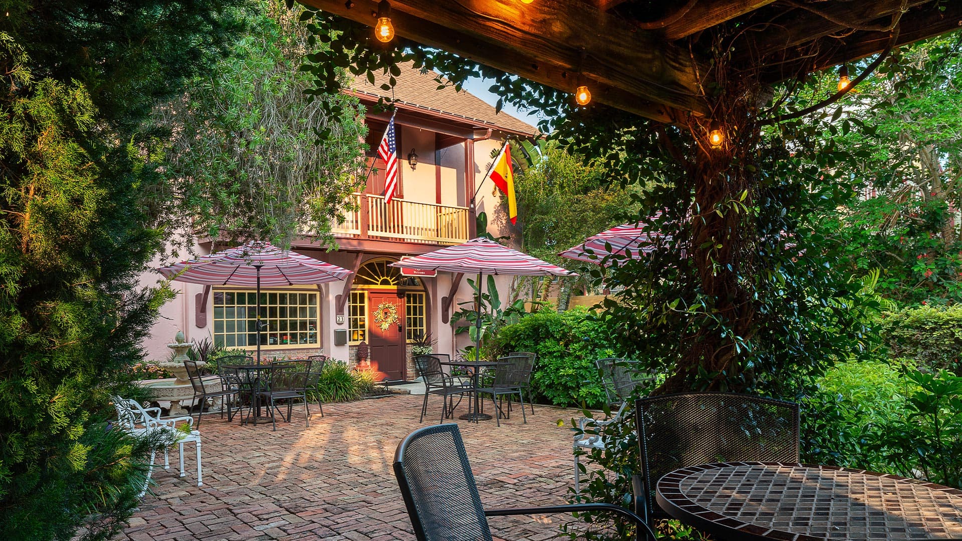 Charming outdoor patio area with tables and umbrellas, framed by lush greenery and a pastel building.