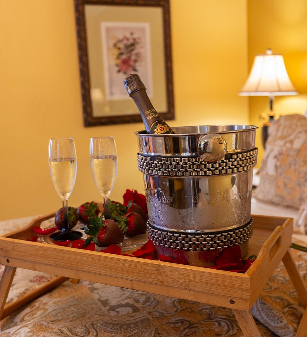 A silver ice bucket with a bottle of champagne, two glasses, strawberries, and rose petals on a wooden tray.