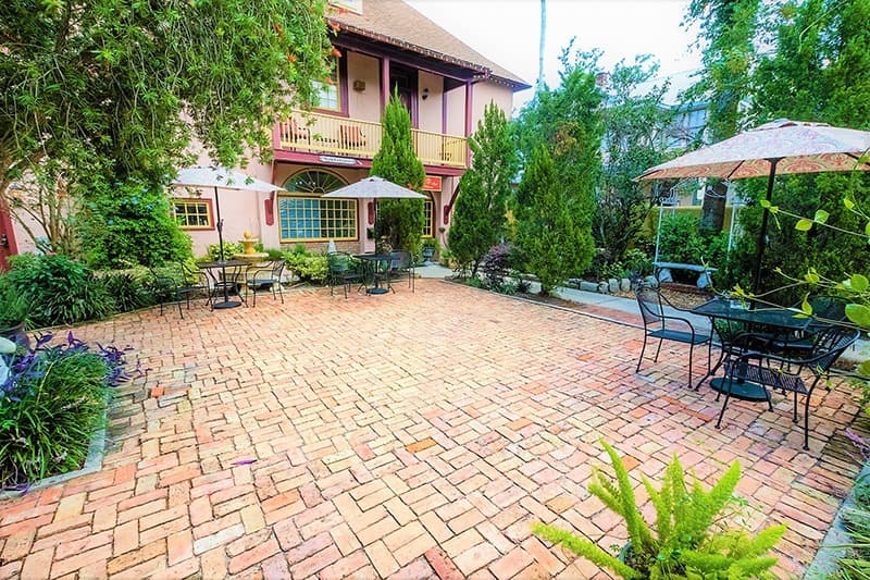 Quaint outdoor patio with brick flooring, tables, and umbrellas surrounded by greenery.
