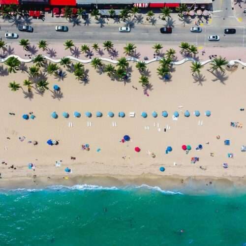 Aerial view of a sandy beach lined with umbrellas, palm trees, and a roadway.